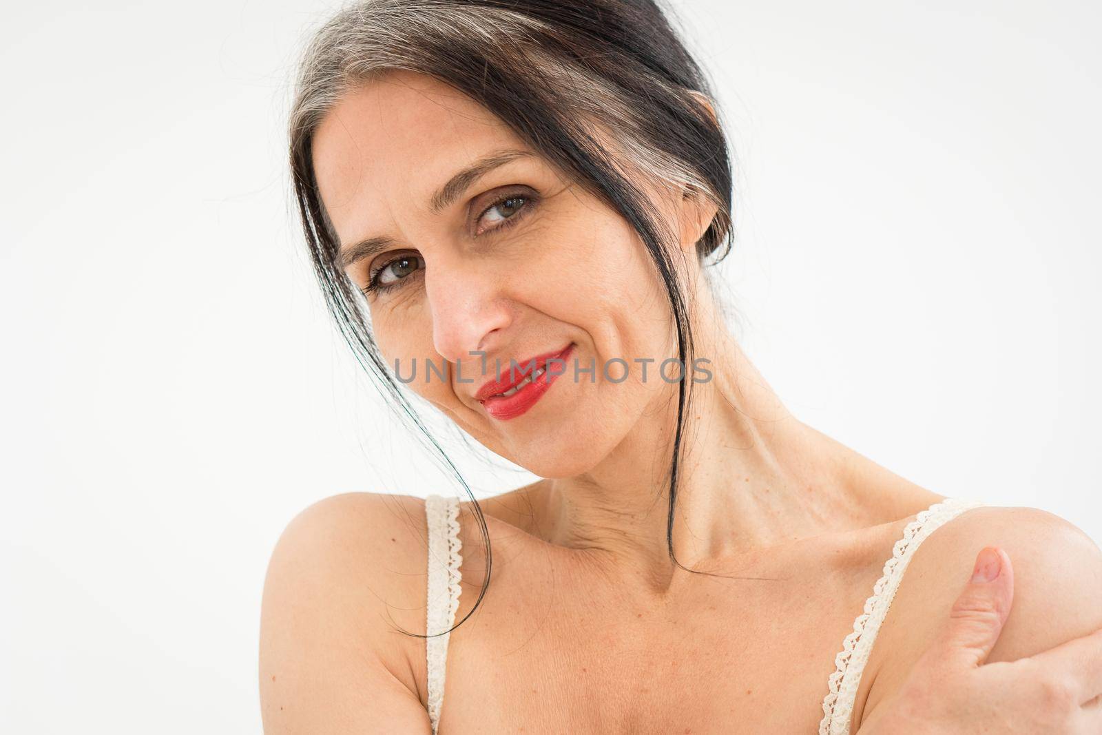 Middle aged woman starting getting grey-haired is posing in studio with naked shoulders on white background, face skin care beauty, middle age skincare cosmetics, cosmetology concept.