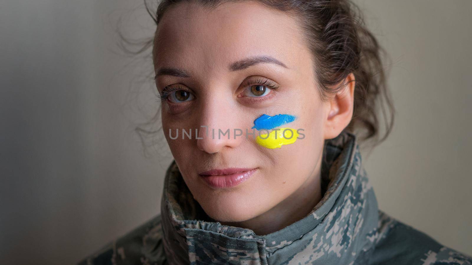 Indoor portrait of young girl with blue and yellow ukrainian flag on her cheek wearing military uniform, mandatory conscription in Ukraine, equality concepts.