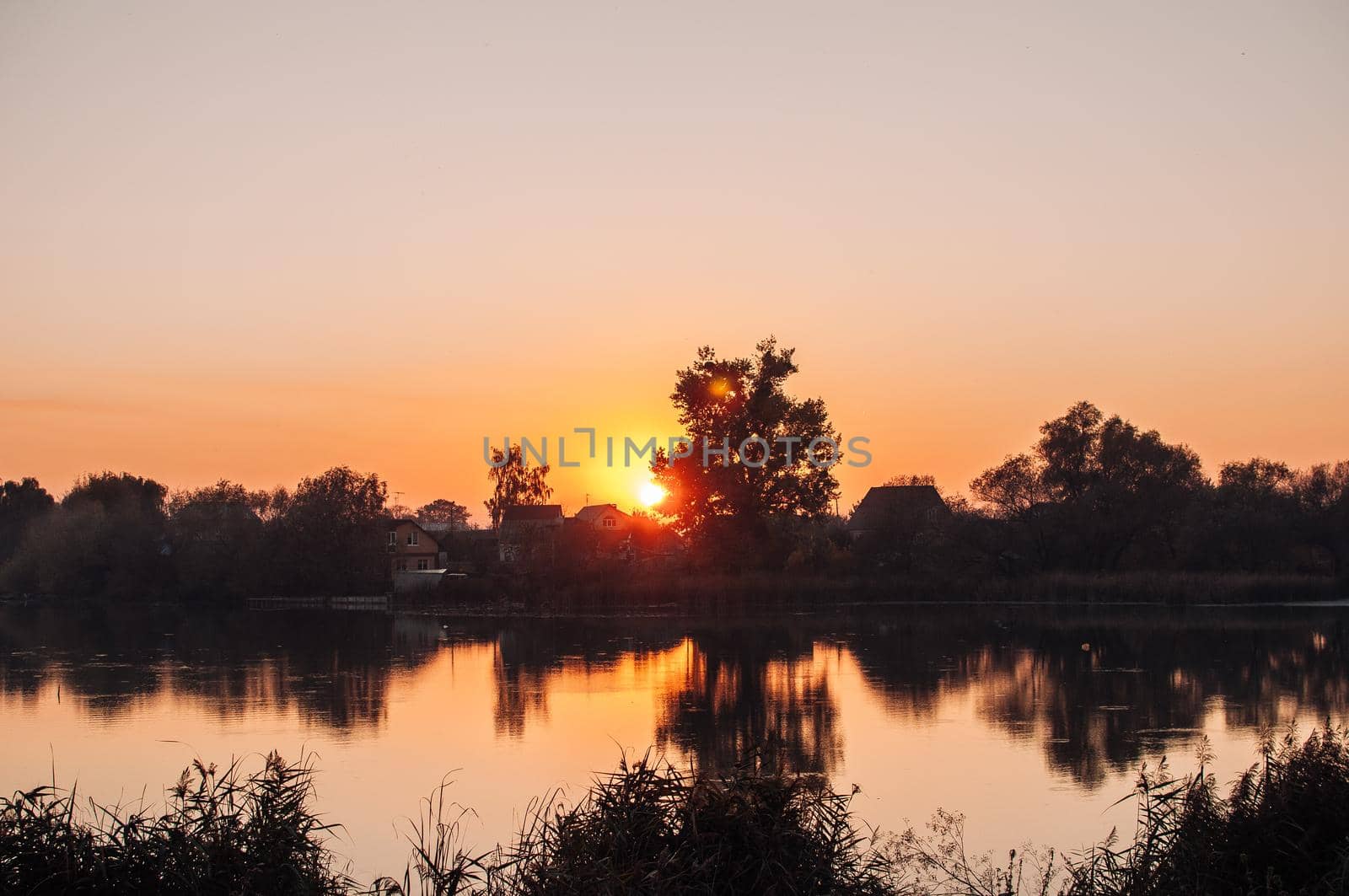 Orange sunset against the background of the river in September in the harvest season. Reflection in the water from the trees. Beautiful autumn landscape. Autumn colors are reflected in calm waters.