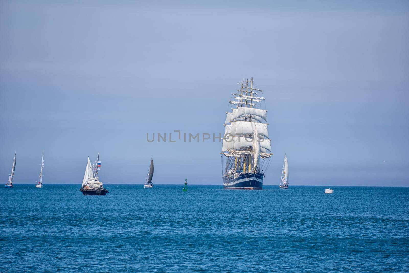 A large ship with white sails sailing on blue water. Nearby are several sailing boats with white sails