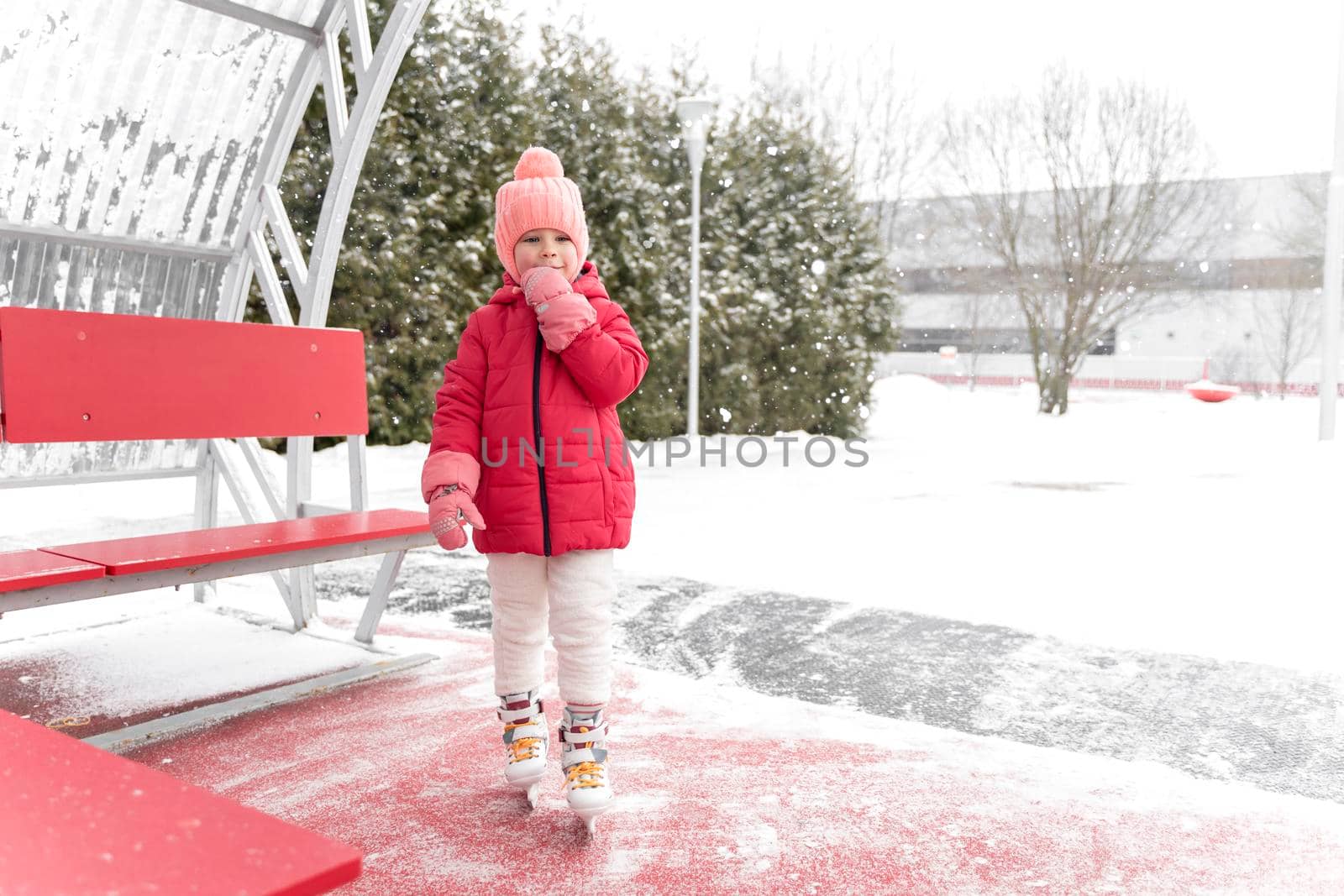 adorable kid girl in a red jacket and skates goes to skate on the ice rink