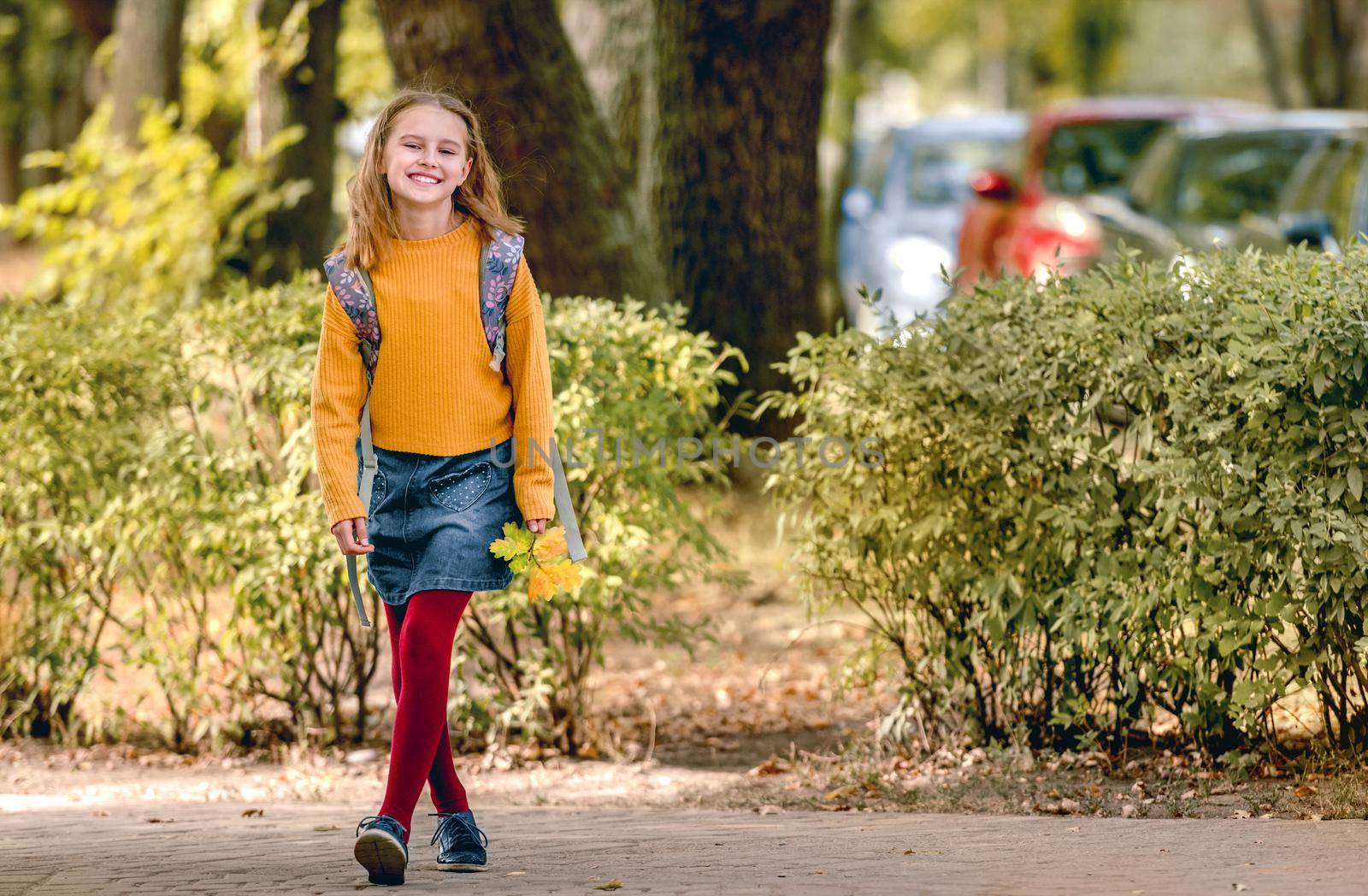 Preteen school girl with backpack holding yellow leaves in her hand, looking at camera and smiling. Pretty pupil female kid after class in autumn park
