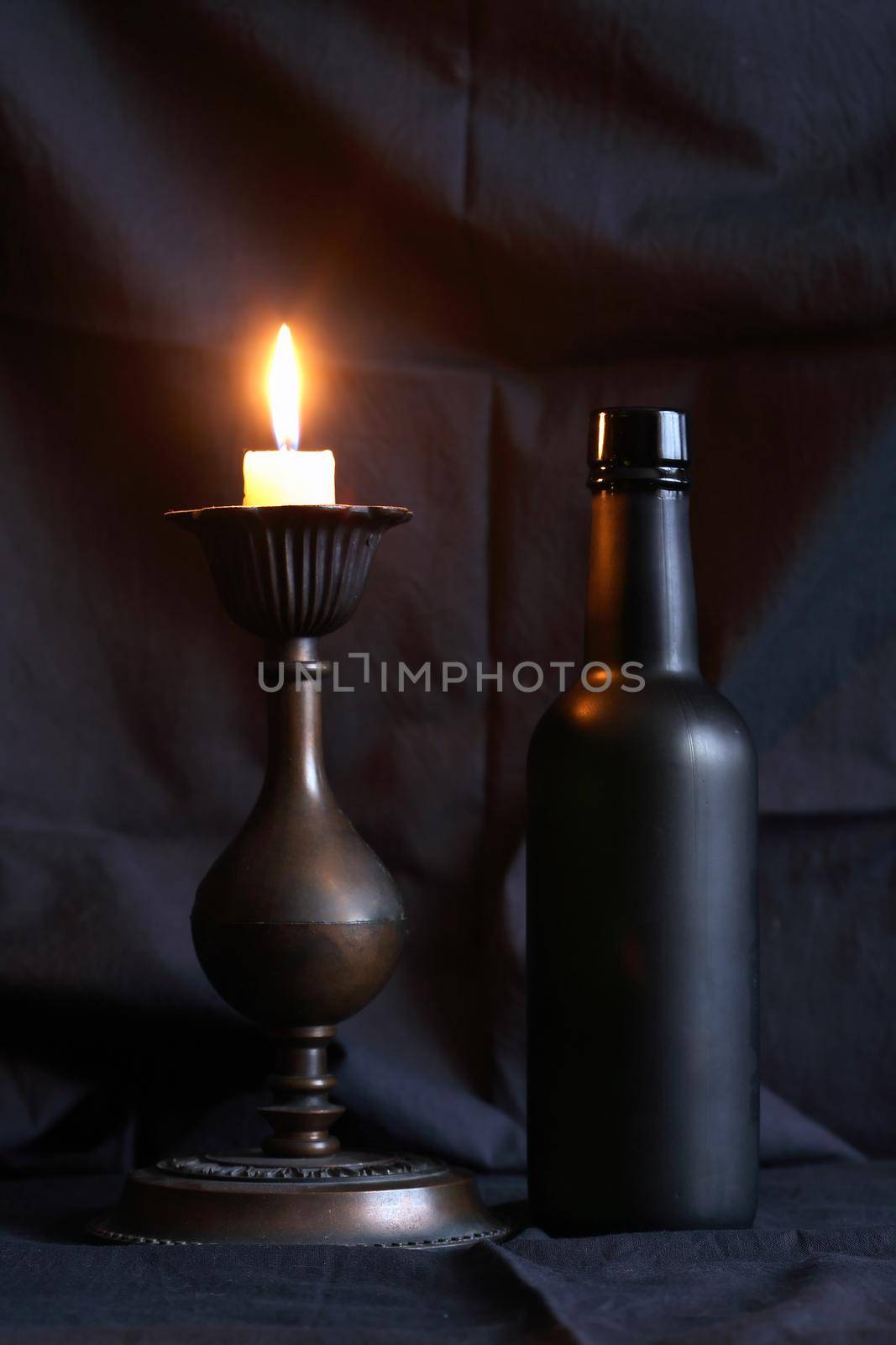 Glass black bottle near lighting candle against nice dark textile background