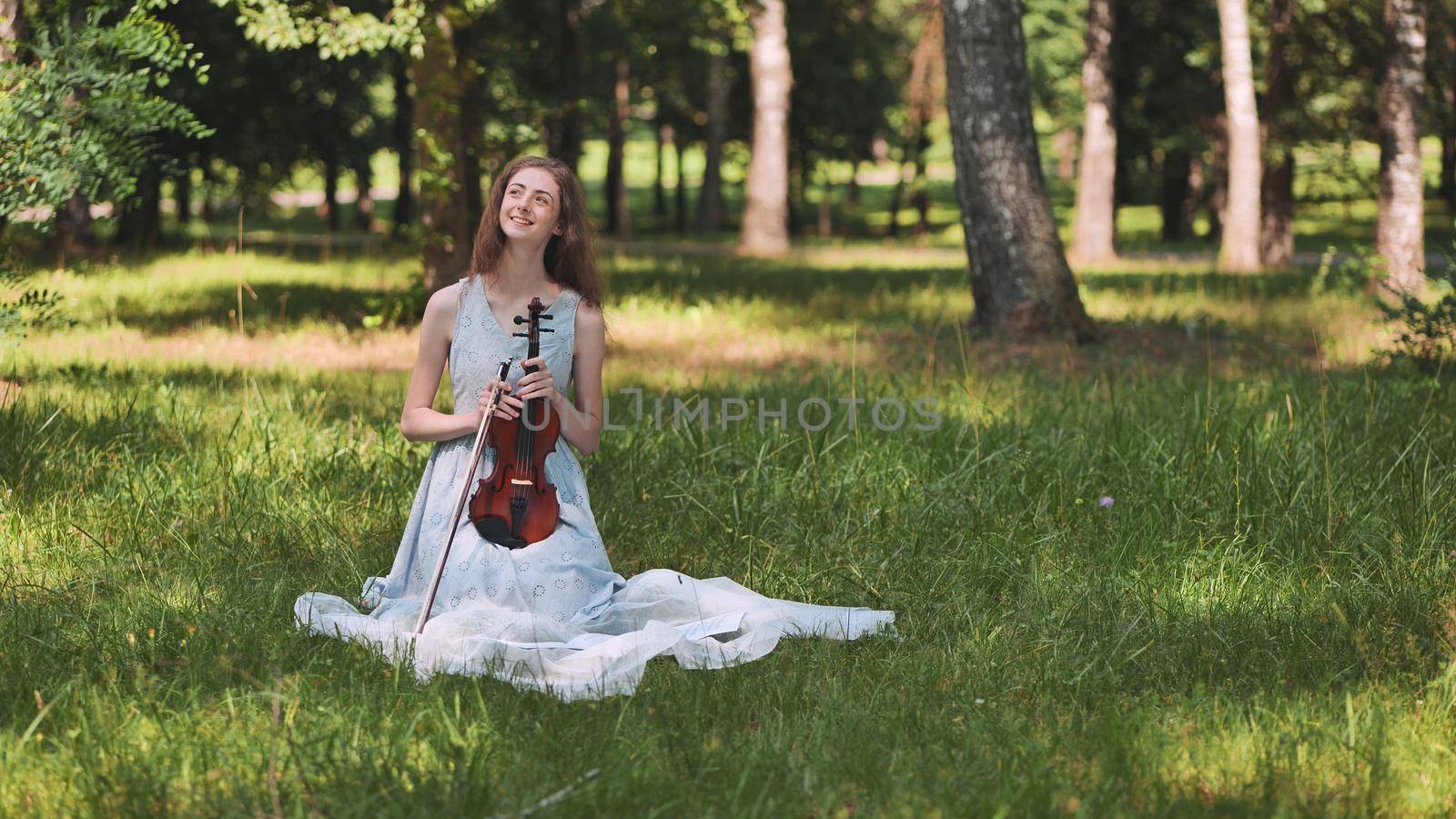 A girl sits with a violin on the grass in a city park