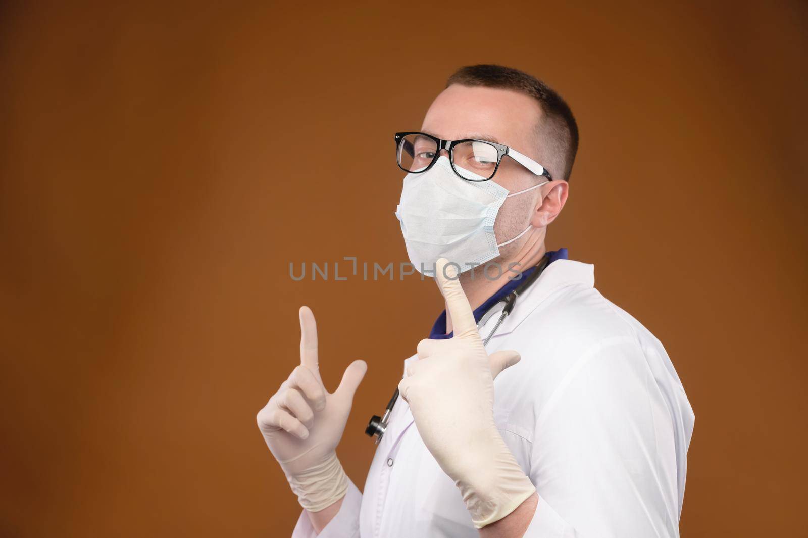 A happy caucasian medic in a white coat with glasses and a mask is dancing in the studio pointing up his index fingers