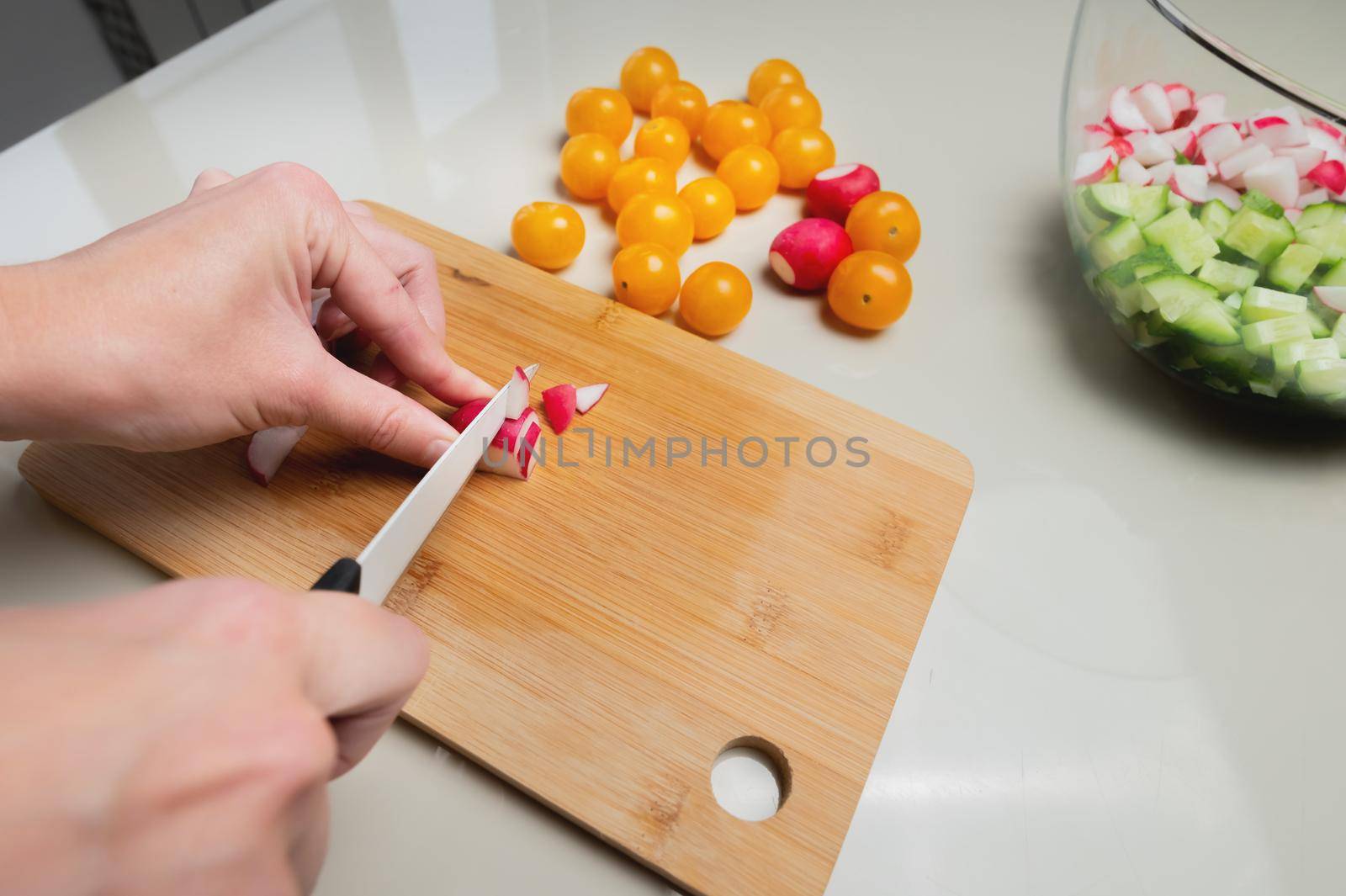 Close-up of female hands cut fresh raw radish on a wooden board against the background of yellow cherry tomatoes. Vegetarian tasty and healthy food.