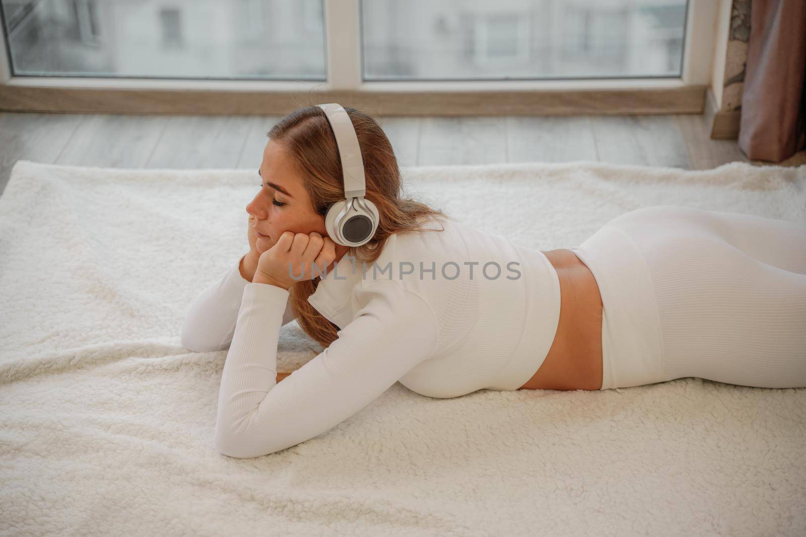 Side view portrait of relaxed woman listening to music with headphones lying on carpet at home. She is dressed in a white tracksuit. by Matiunina