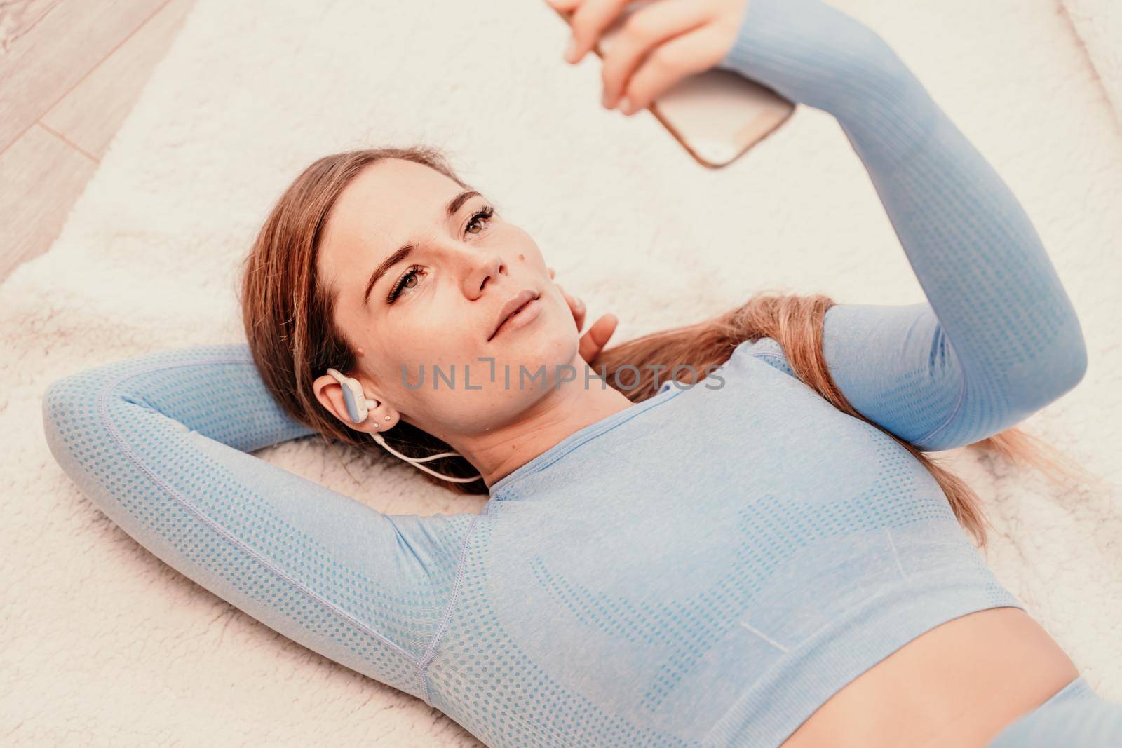 Top view portrait of relaxed woman listening to music with headphones lying on carpet at home. She is dressed in a blue tracksuit. by Matiunina