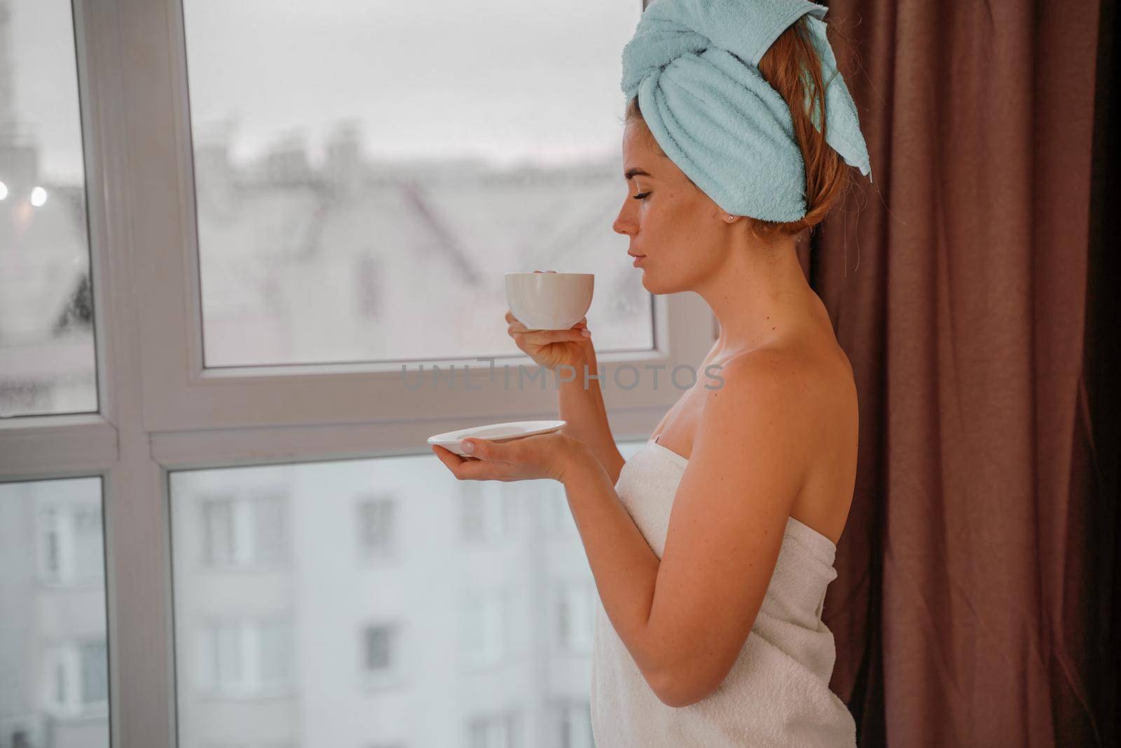 Young serene relaxed woman in spa bath towel drinking hot beverage tea coffee after taking shower bath at home. Beauty treatment, hydration concept