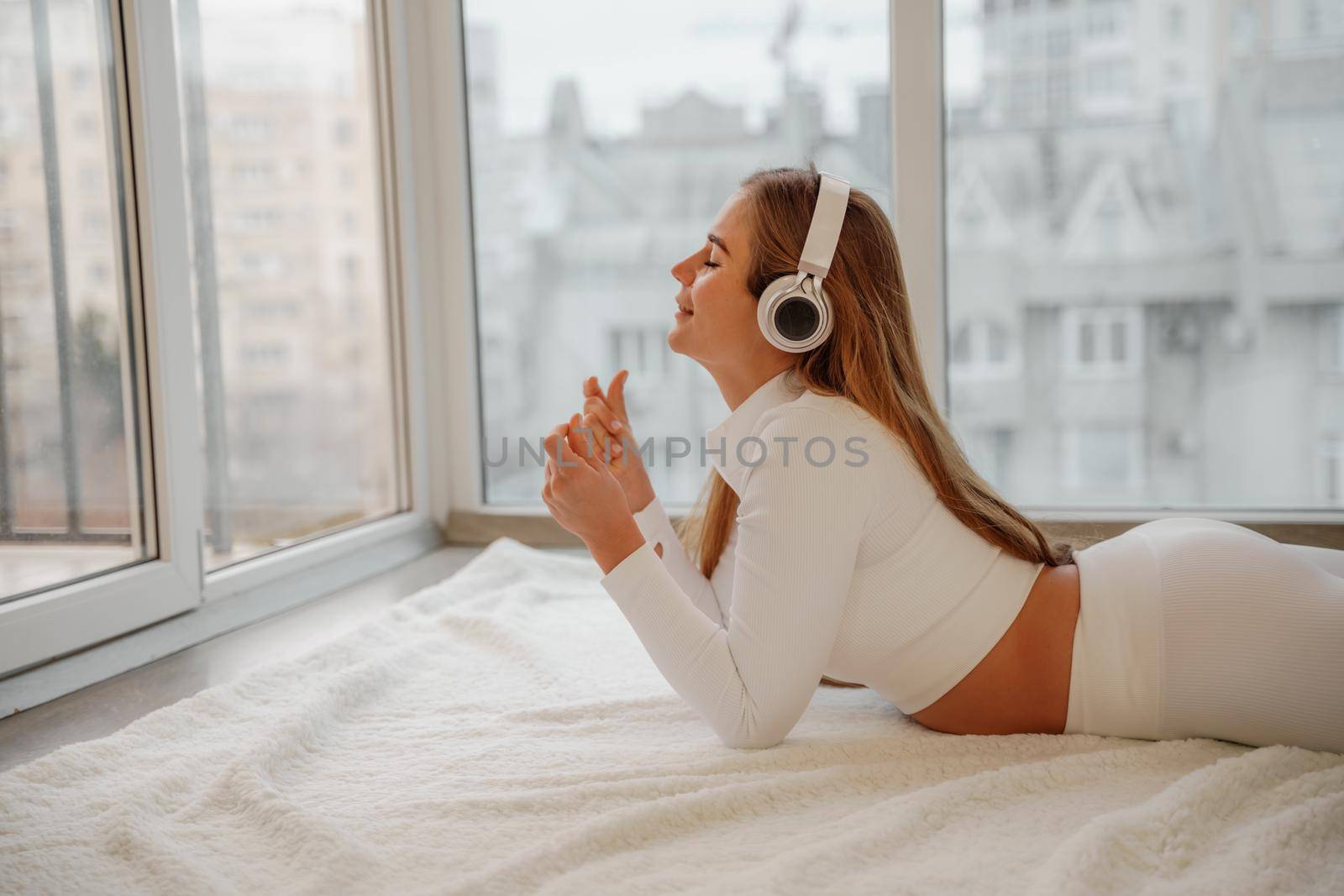 Side view portrait of relaxed woman listening to music with headphones lying on carpet at home. She is dressed in a white tracksuit