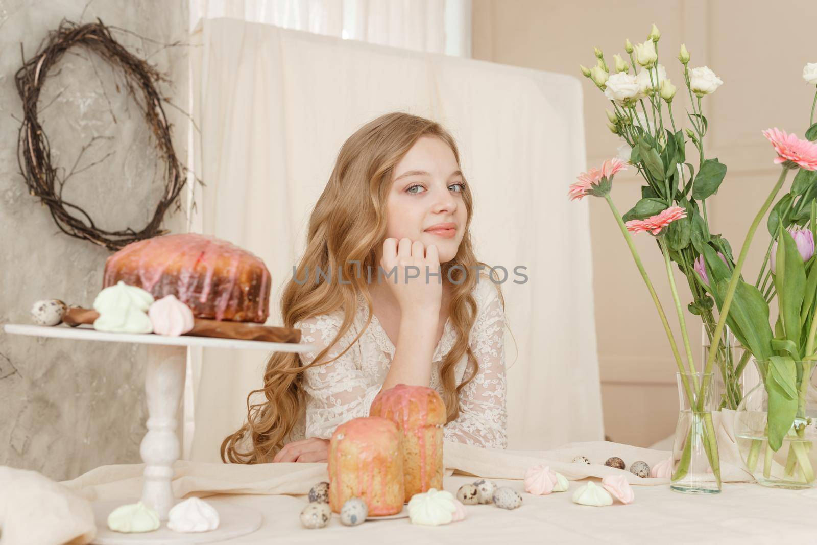 A girl with long hair in a light dress is sitting at the Easter table with cakes, spring flowers and quail eggs. Happy Easter celebration