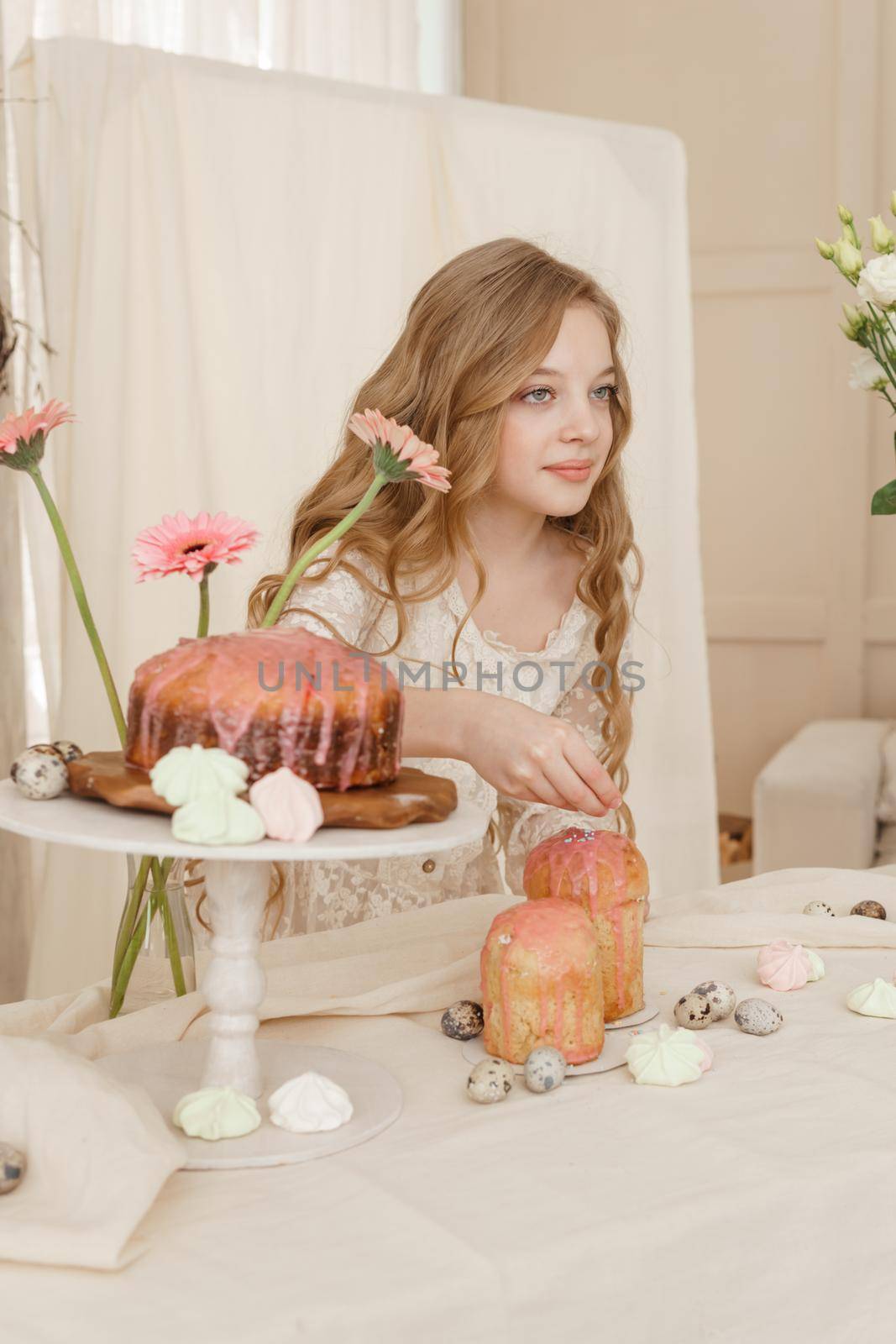 A girl with long hair in a light dress is sitting at the Easter table with cakes, spring flowers and quail eggs. Happy Easter celebration