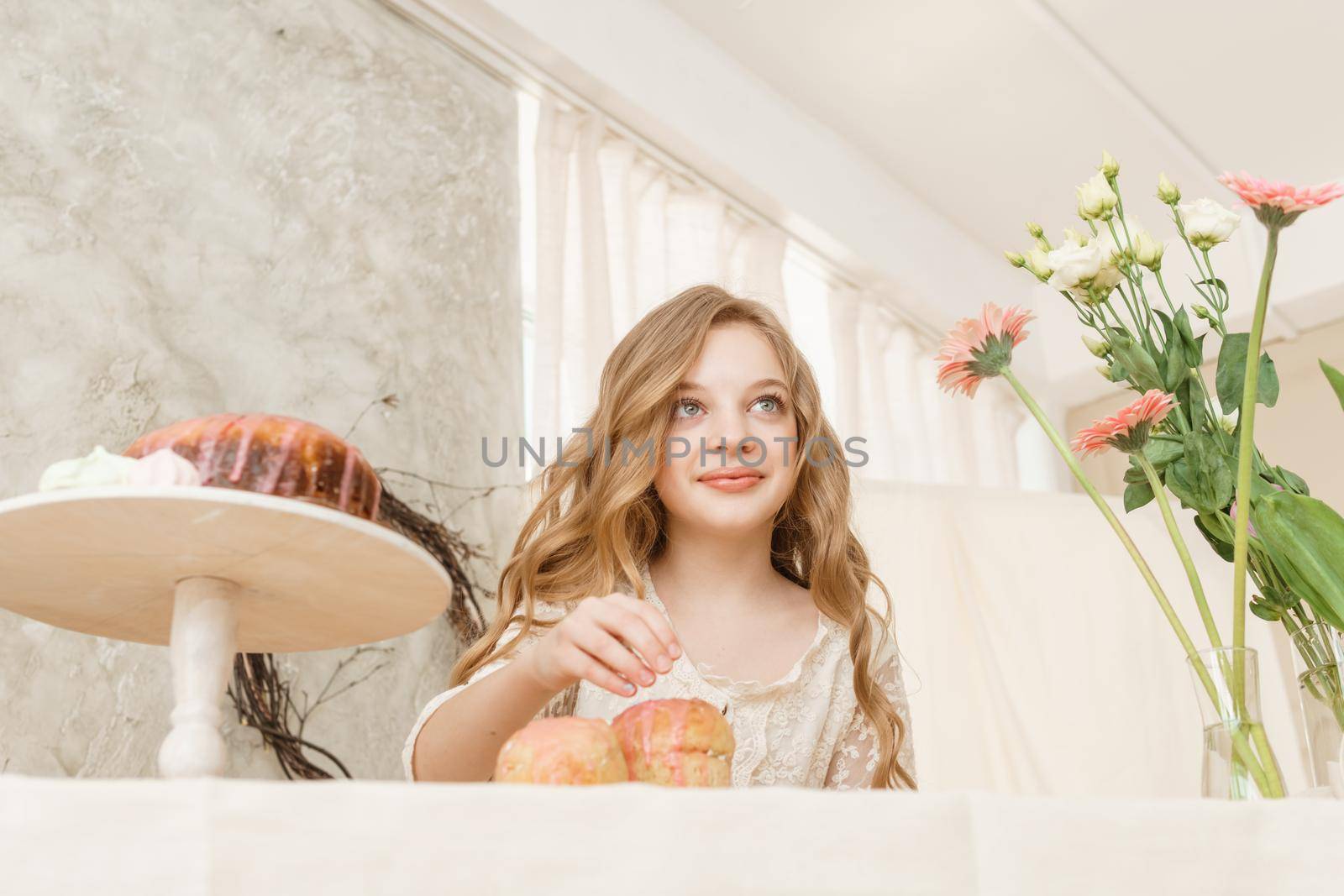 A girl with long hair in a light dress is sitting at the Easter table with cakes, spring flowers and quail eggs. Happy Easter celebration