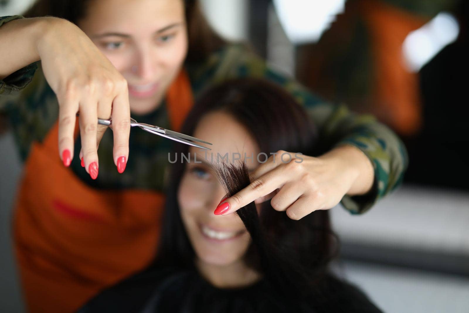 Portrait of concentrated professional hairdresser hold lock of hair and cutting with scissors. Smiling female client sit in masters chair. Wellness concept