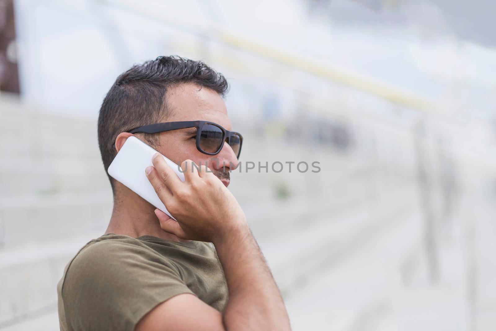Happy male in sunglasses standing outdoors, talking by smartphone