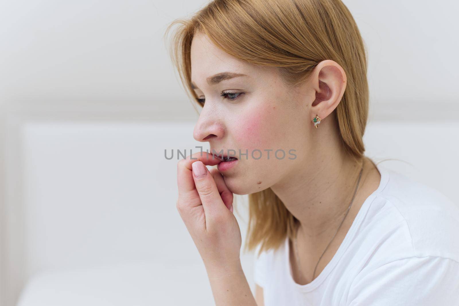 A young woman sits in bed, worries and bites her nails. Worried casual woman sitting on her bed in bedroom.