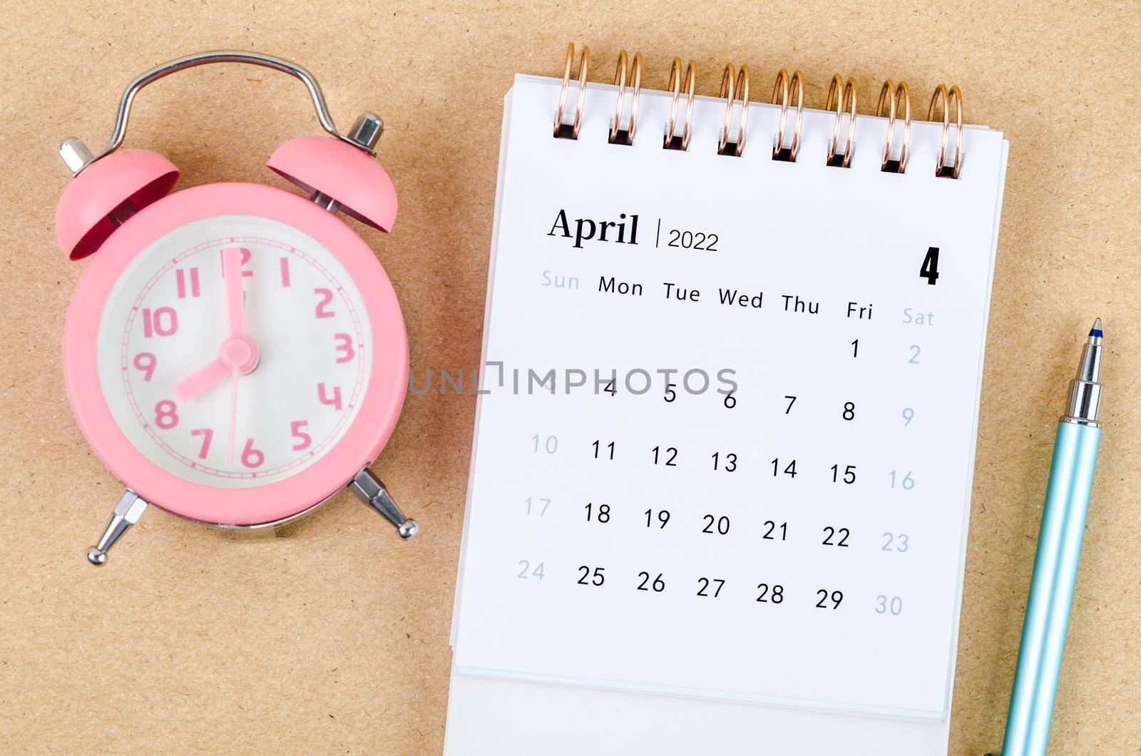 April 2022 Desk calendar with pink alarm clock on brown background.