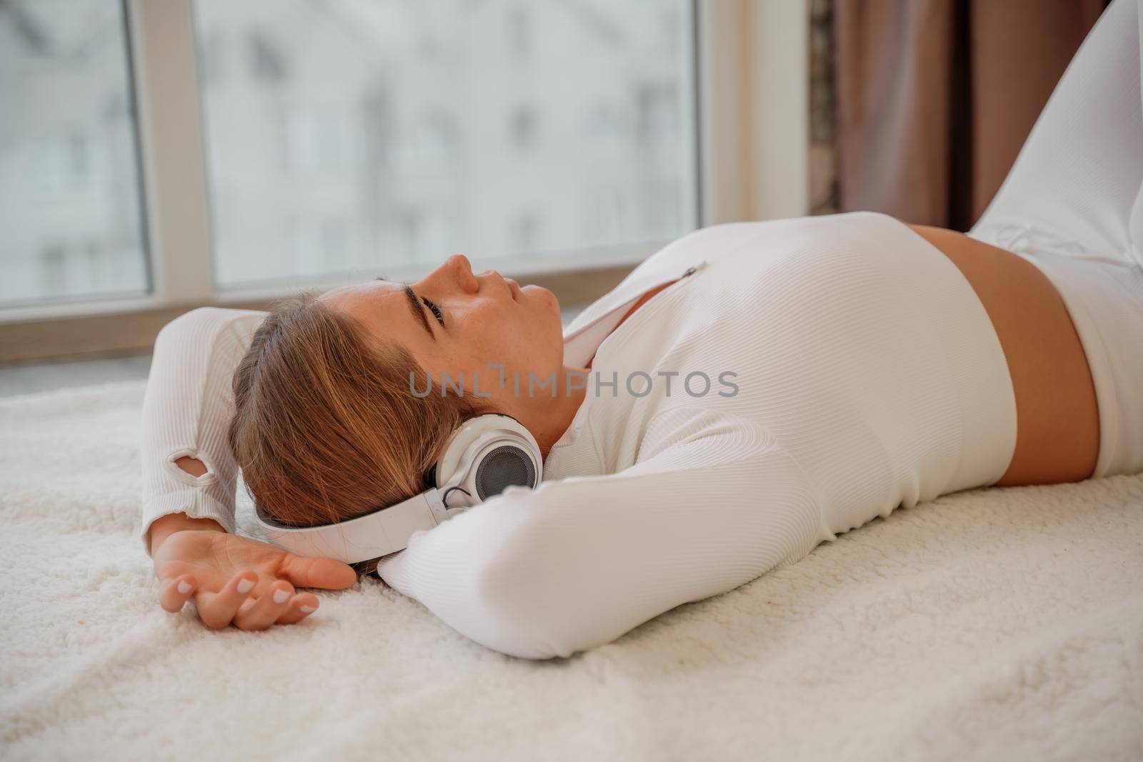 Side view portrait of relaxed woman listening to music with headphones lying on carpet at home. She is dressed in a white tracksuit. by Matiunina