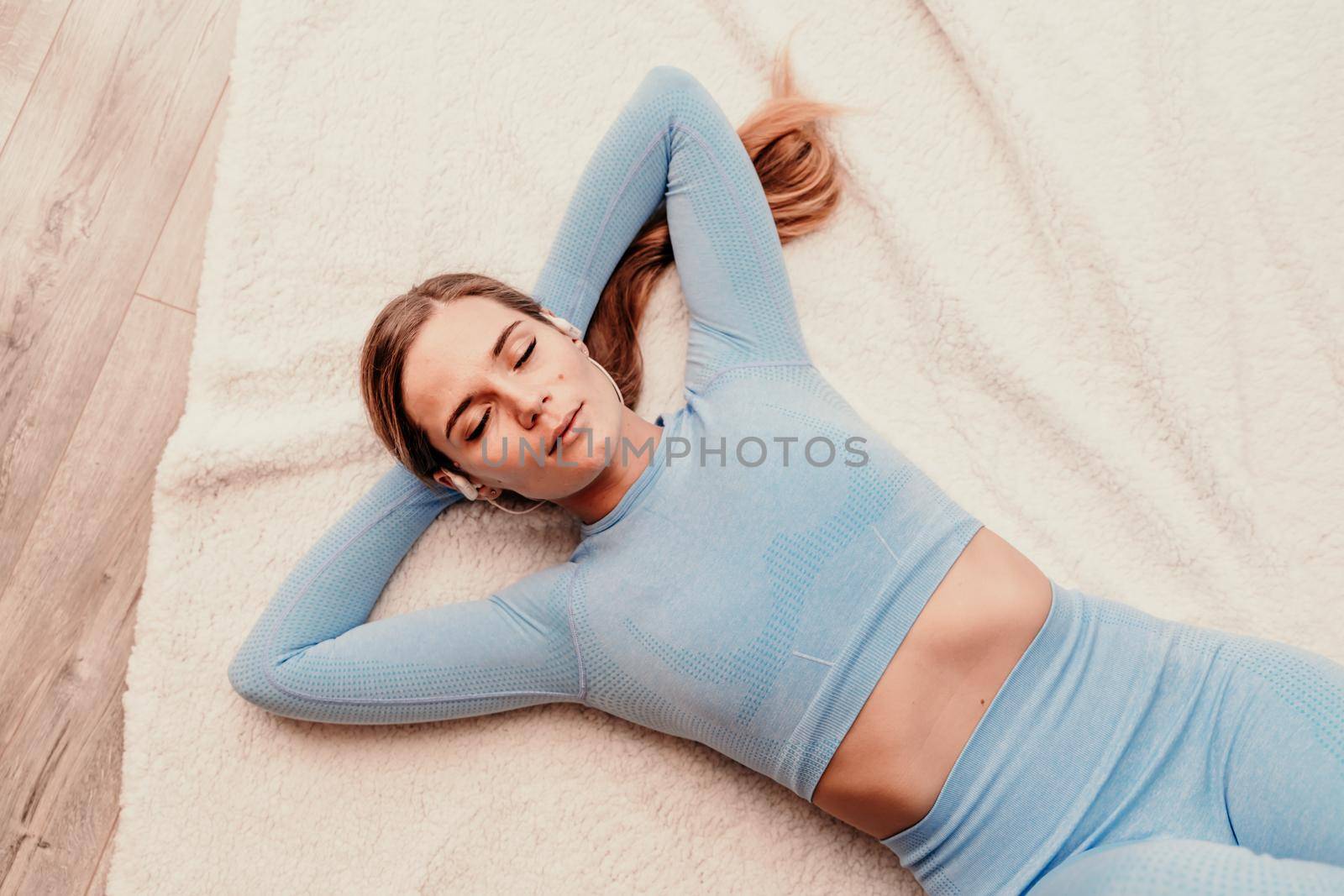 Top view portrait of relaxed woman listening to music with headphones lying on carpet at home. She is dressed in a blue tracksuit. by Matiunina