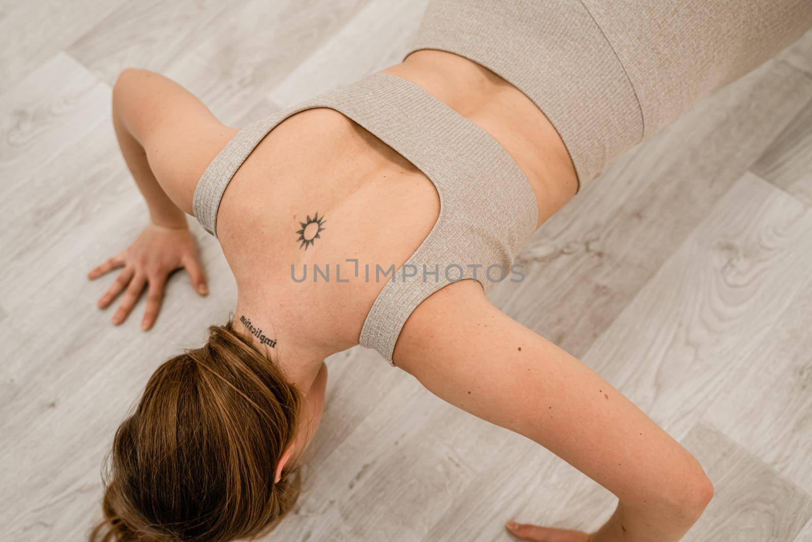 Top view of a muscular woman during push-ups. She is wearing a beige top and leggings, with a round tattoo between her shoulder blades. The concept of a healthy lifestyle. by Matiunina