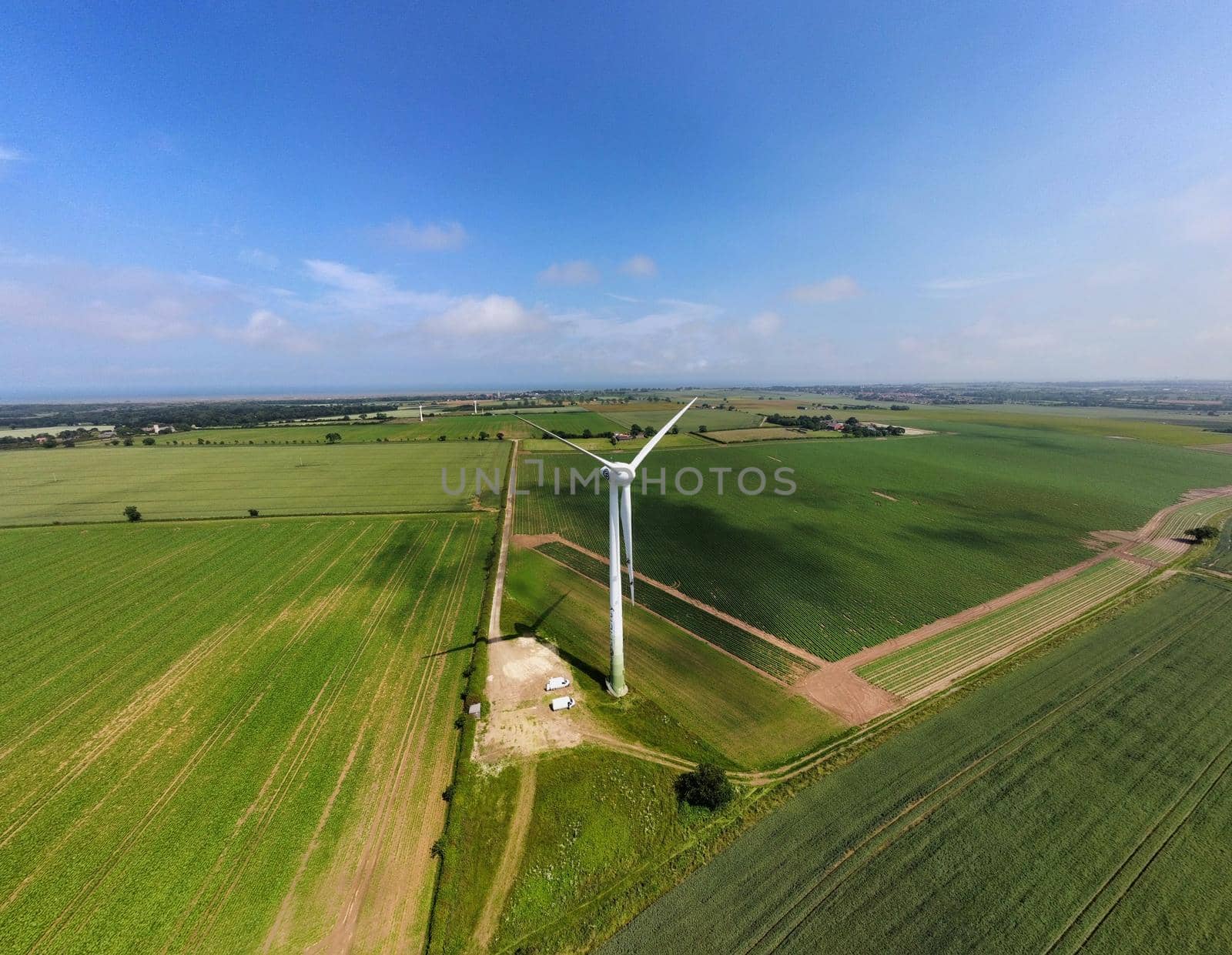 Drone shot of a single wind turbine up close, standing tall amongst a patchwork of green fields with prominent shadows from clouds overhead. Taken on a summer day in England, UK.
