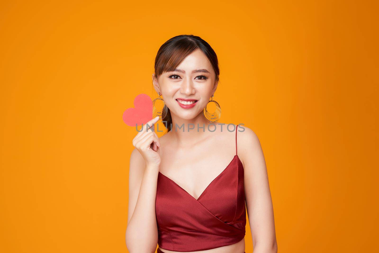 happy young woman dressed in red dress holding paper heart isolated over yellow background