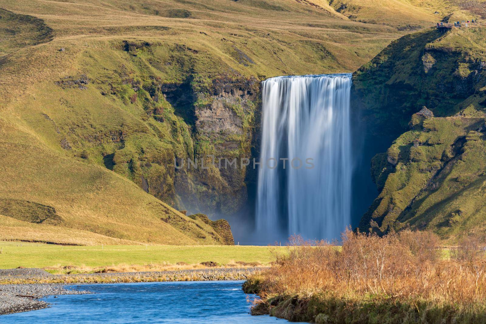 Long exposure of famous Skogafoss waterfall from the distance with hikers on top viewpoint by FerradalFCG