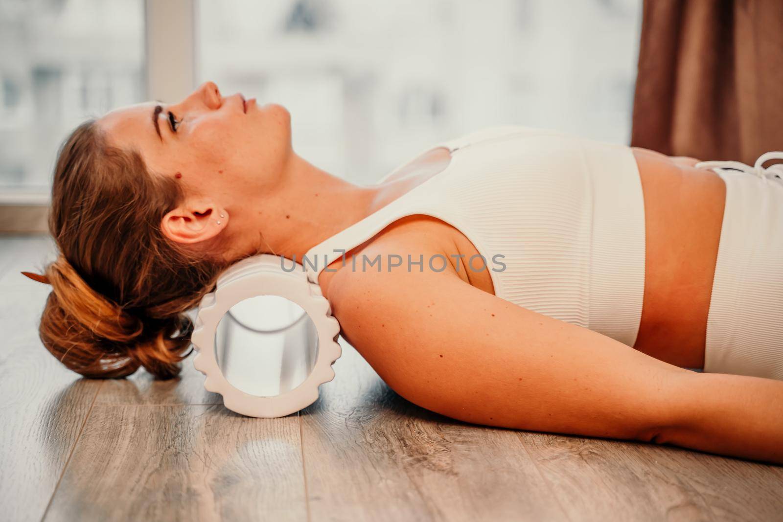 Adult athletic woman, in white bodysuit, performing fascia exercises on the floor - caucasian woman using a massage foam roller - a tool to relieve tension in the back and relieve muscle pain - the concept of physiotherapy and stretching training.