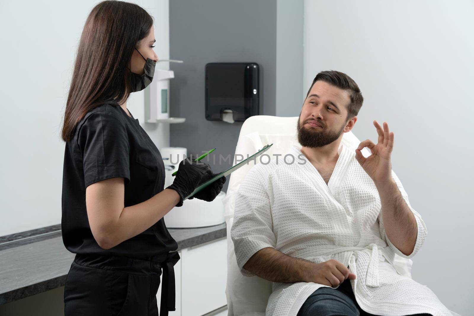 Expert female cosmetologist giving consultation to young man patient sitting on couch in beautician office of aesthetic clinic