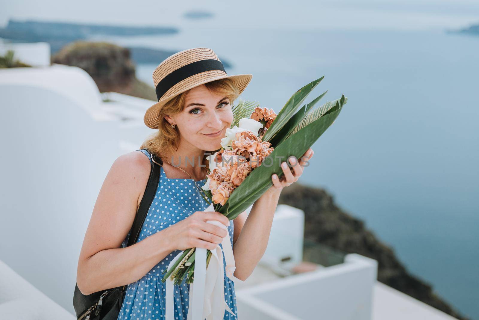 Beautiful girl walking with bouquet of flowers along the street of Santorini island, an old European town, Greece. Portrait of a tourist girl walking on the street background. Love story summer vacation by Andrii_Ko