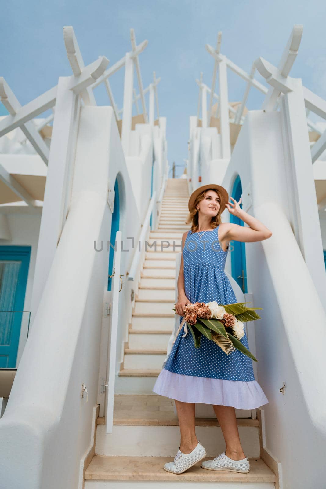 Beautiful girl walking with bouquet of flowers along the street of Santorini island, an old European town, Greece. Portrait of a tourist girl walking on the street background. Love story summer vacation by Andrii_Ko