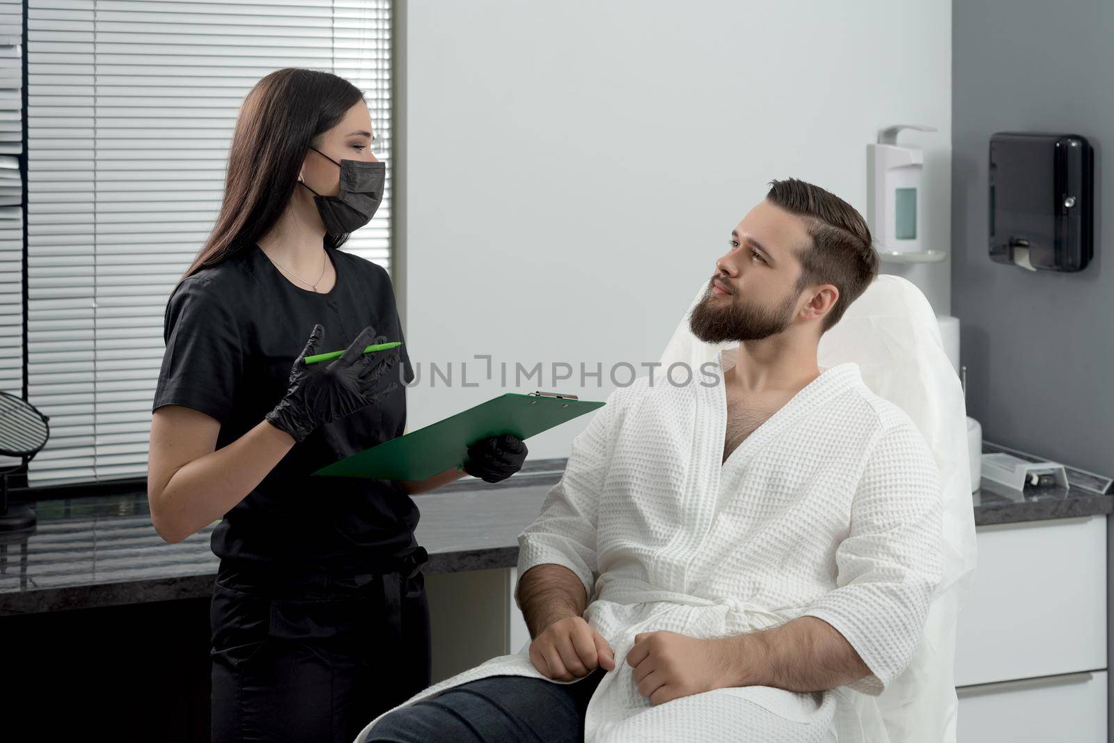 Expert female cosmetologist giving consultation to young man patient sitting on couch in beautician office of aesthetic clinic