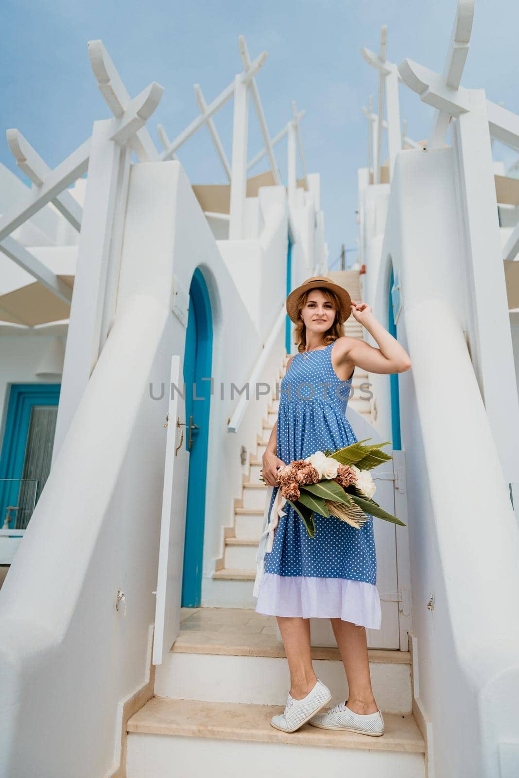 Beautiful girl walking with bouquet of flowers along the street of Santorini island, an old European town, Greece. Portrait of a tourist girl walking on the street background. Love story summer vacation by Andrii_Ko