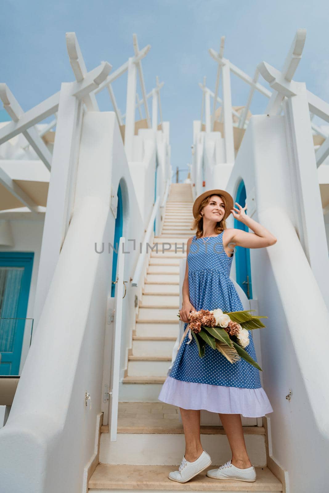 Beautiful girl walking with bouquet of flowers along the street of Santorini island, an old European town, Greece. Portrait of a tourist girl walking on the street background. Love story summer vacation by Andrii_Ko