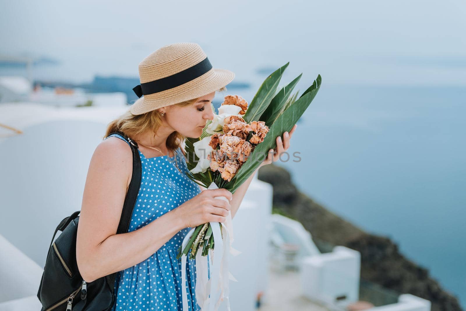 Beautiful girl walking with bouquet of flowers along the street of Santorini island, an old European town, Greece. Portrait of a tourist girl walking on the street background. Love story summer vacation by Andrii_Ko