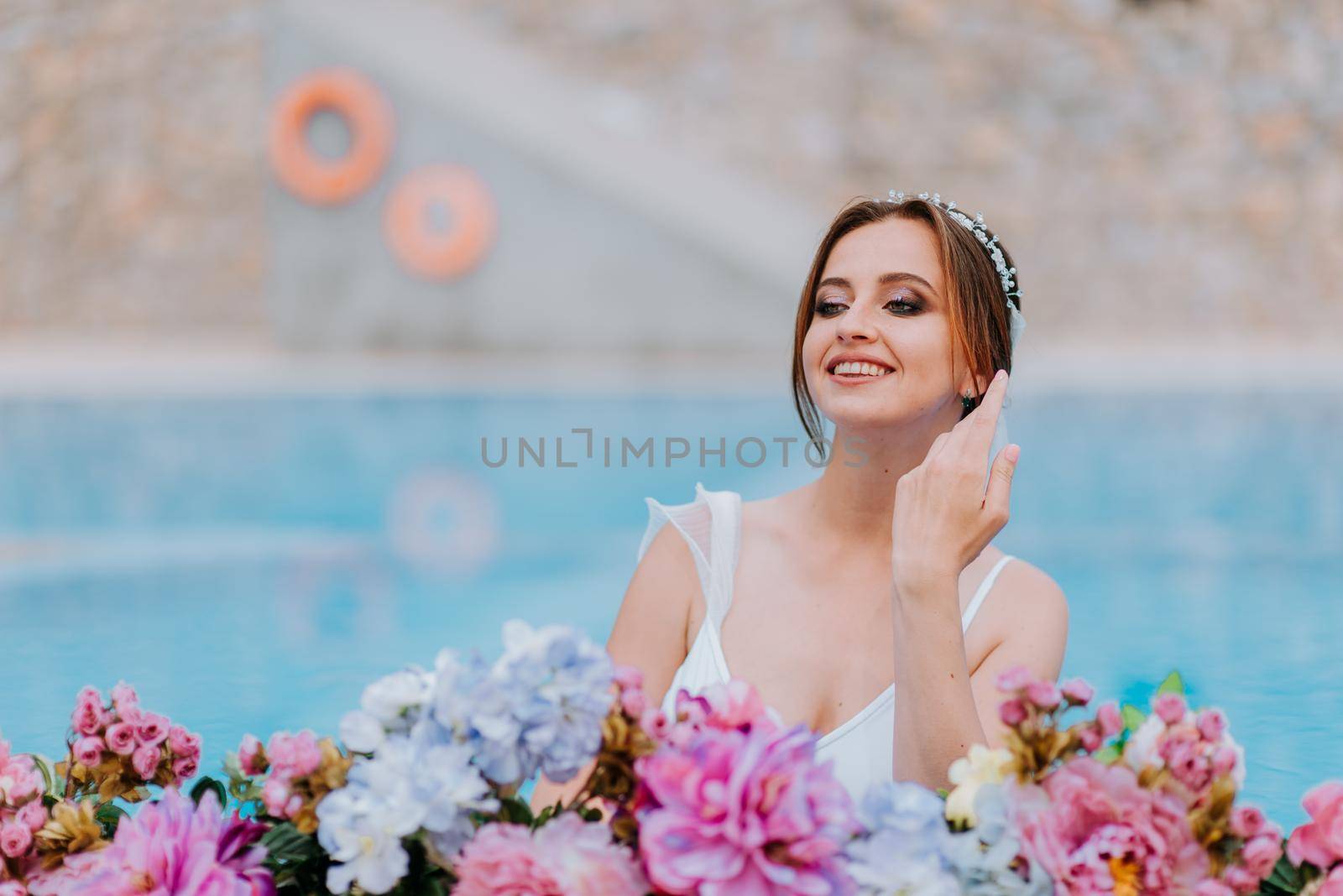 Beautiful bride in a beautiful white swimsuit and garlands of flowers, standing in swimming pool with flower on turquoise water, beautiful girl in water pool in sunny day.