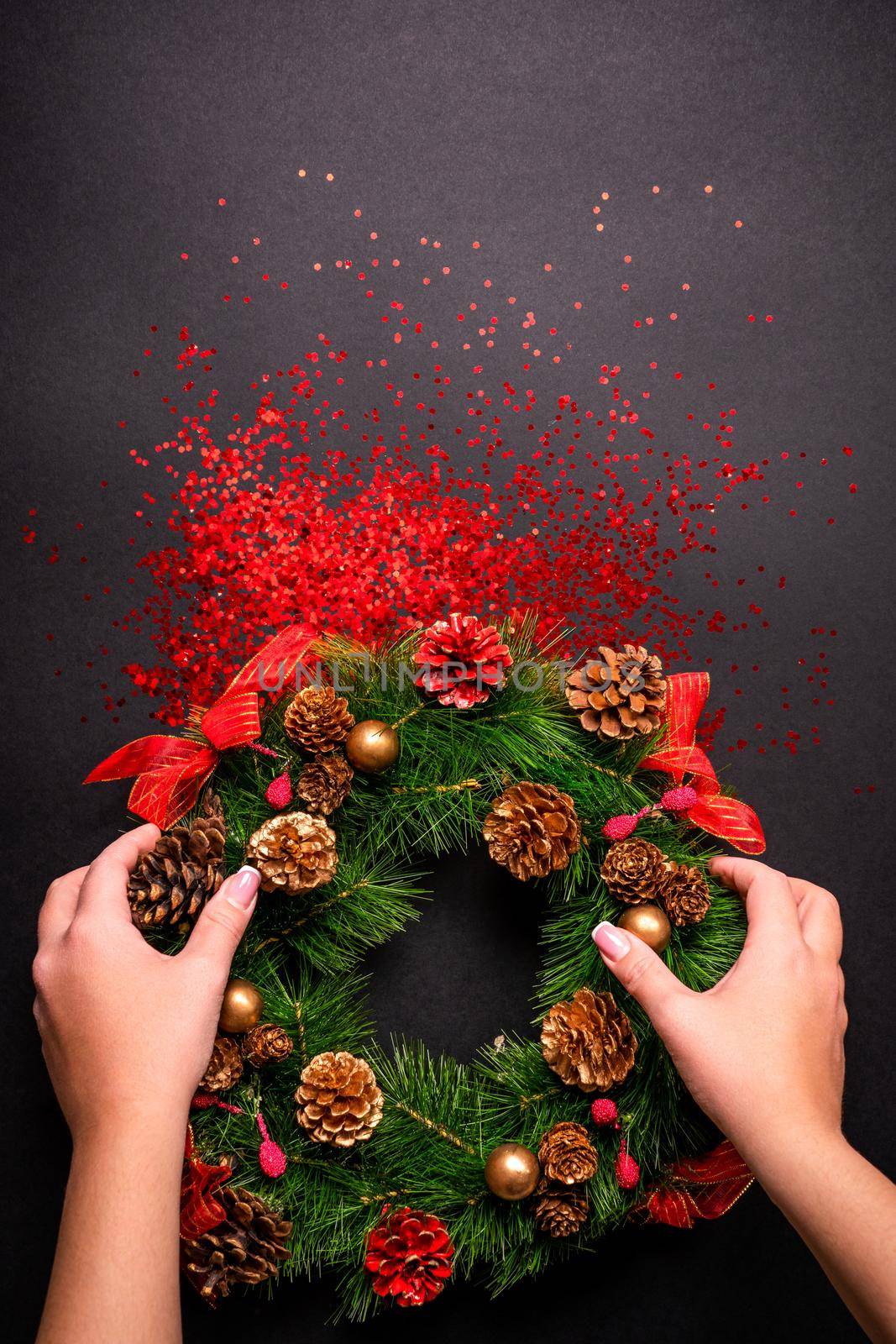 Female hands holding christmas wreath over black background with glitter and copy space, top view