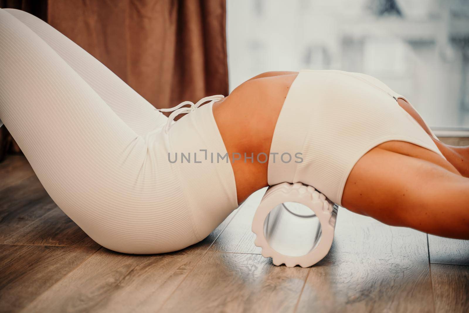 Adult athletic woman, in white bodysuit, performing fascia exercises on the floor - caucasian woman using a massage foam roller - a tool to relieve tension in the back and relieve muscle pain - the concept of physiotherapy and stretching training by Matiunina