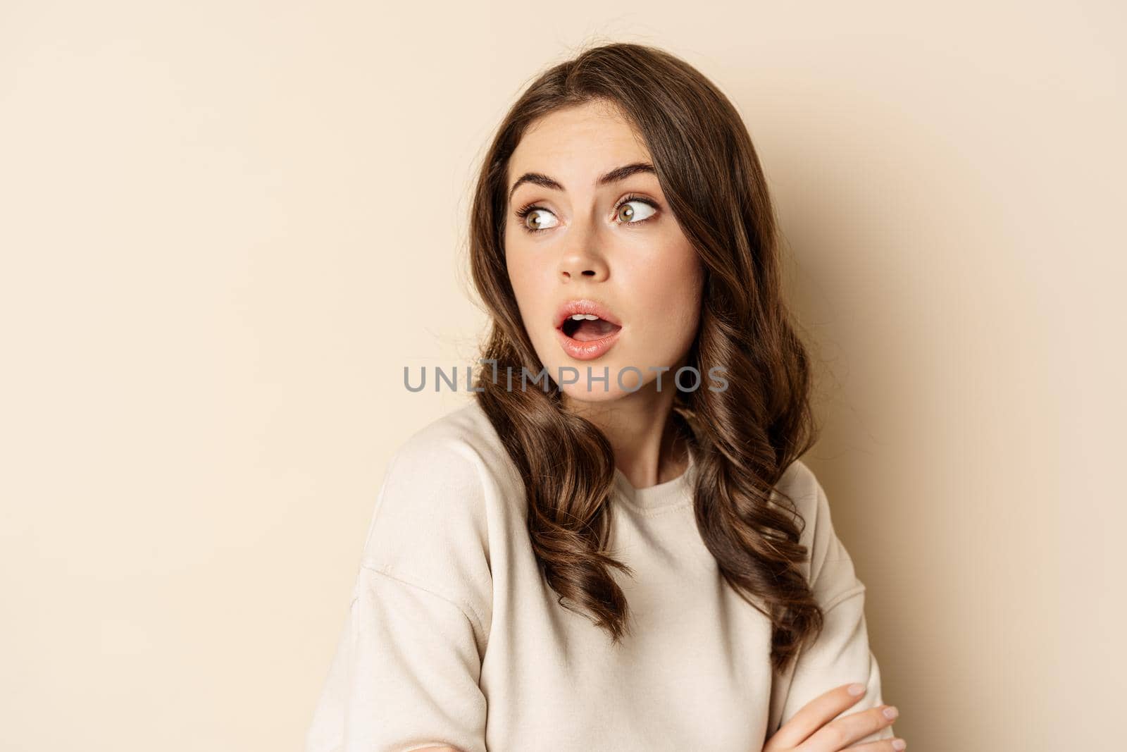 Close up portrait of brunette feminine woman turn back, look behind with surprised face expression, standing against beige background.