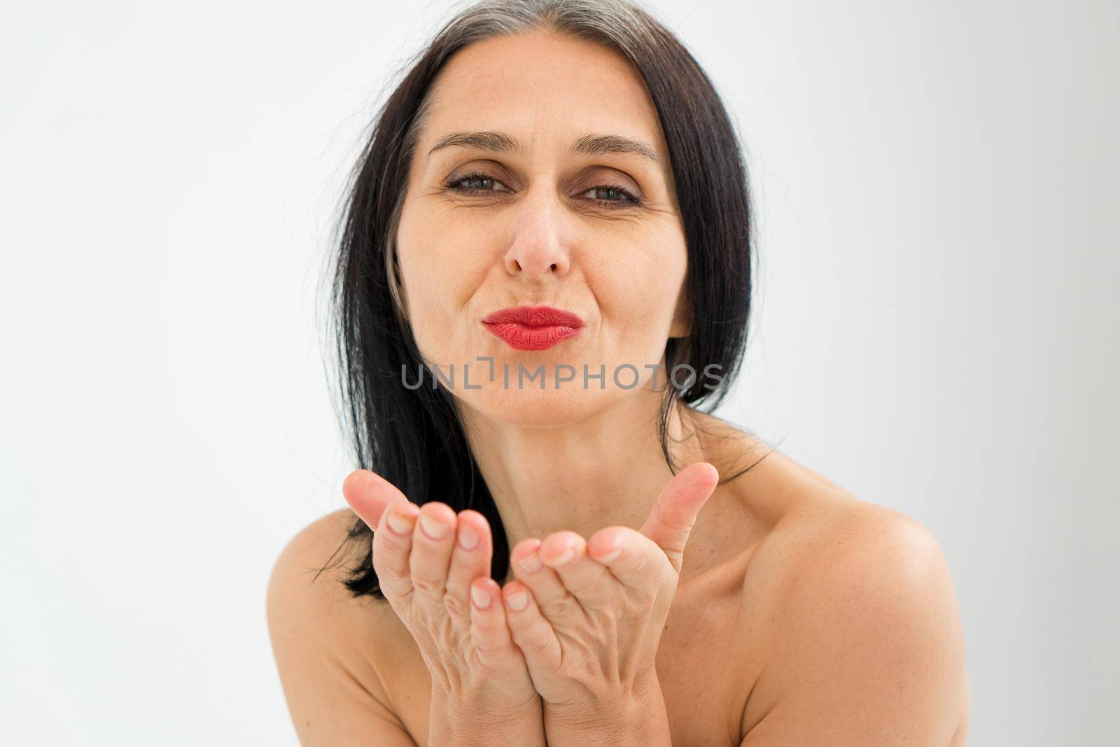 Middle aged woman starting getting grey-haired is sending air kiss in studio with naked shoulders on white background, face skin care beauty, middle age skincare cosmetics, cosmetology concept.