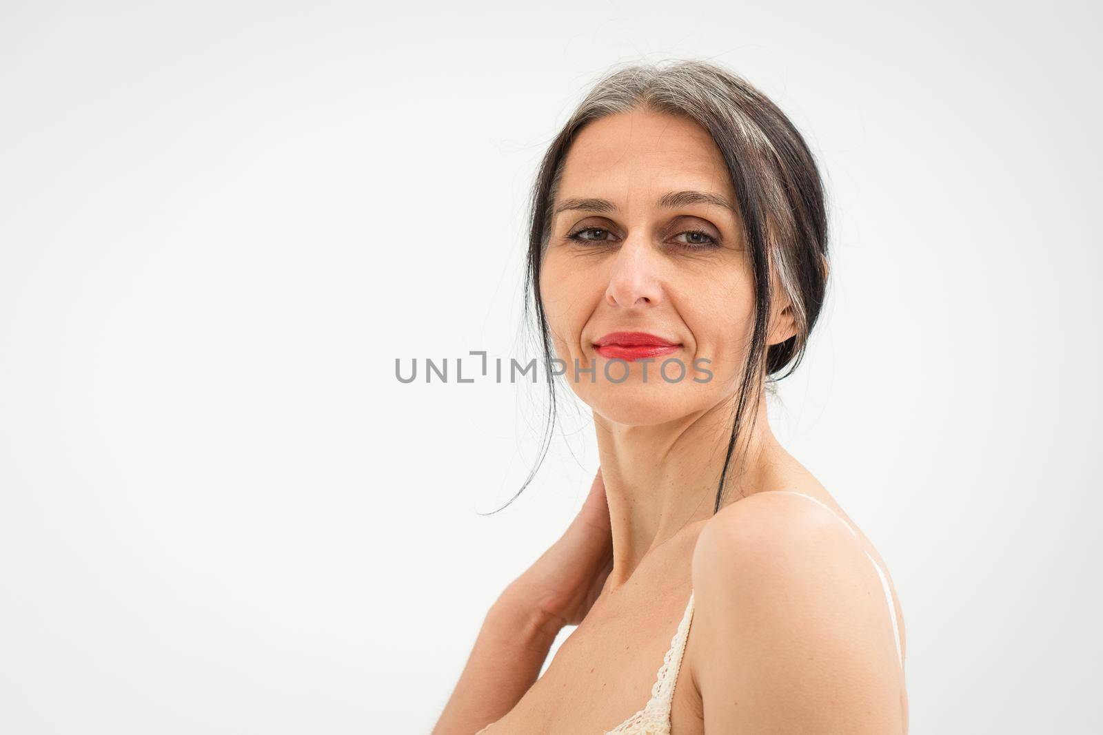 Middle aged woman starting getting grey-haired is posing in studio with naked shoulders on white background, face skin care beauty, middle age skincare cosmetics, cosmetology concept.