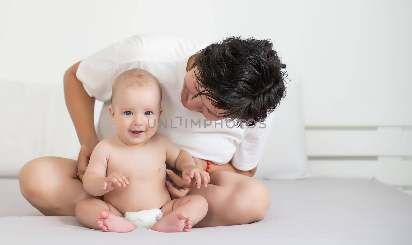 Mother and child on a white bed. Mom and baby boy in diaper playing in bedroom. Parent and little kid relaxing at home. Family having fun together