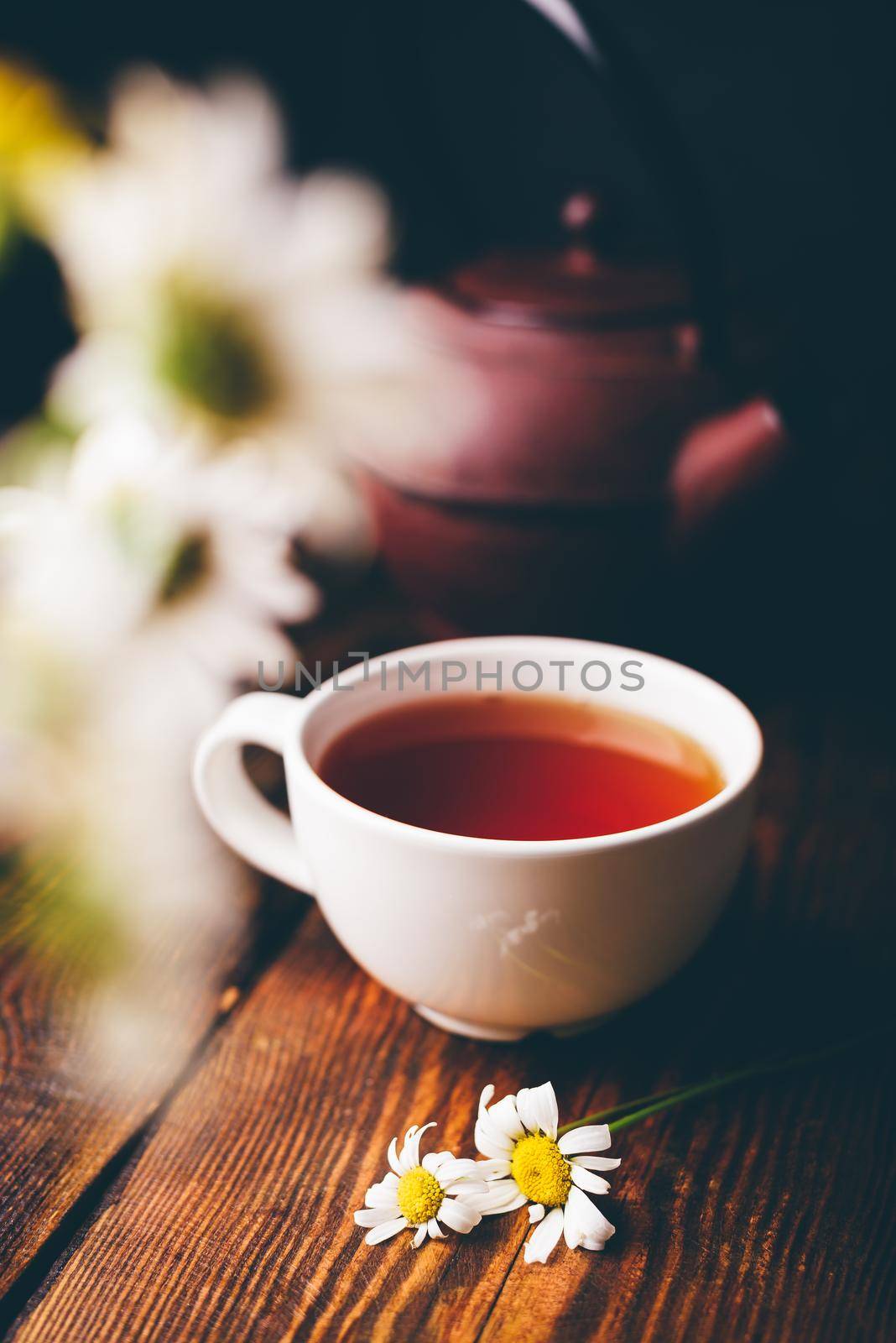 White cup of tea and chamomile flowers on wooden table. Blurred bouquet on foreground