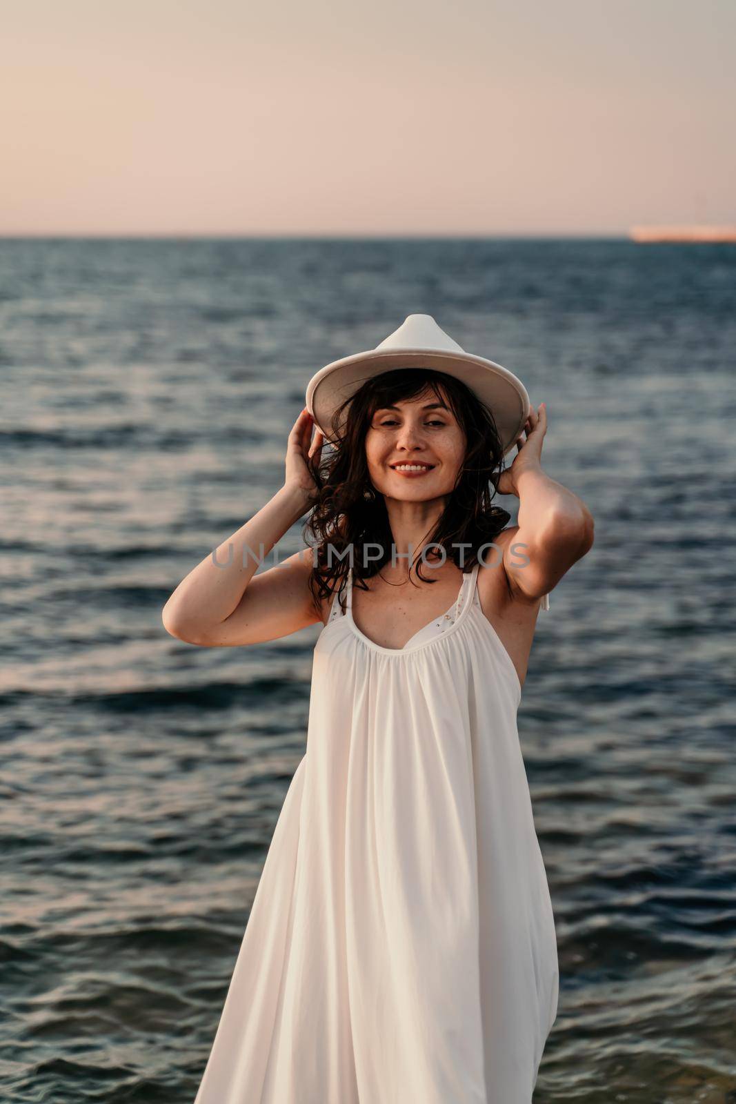 A woman in a white dress and hat is standing on the beach enjoying the sea. Happy summer holidays.