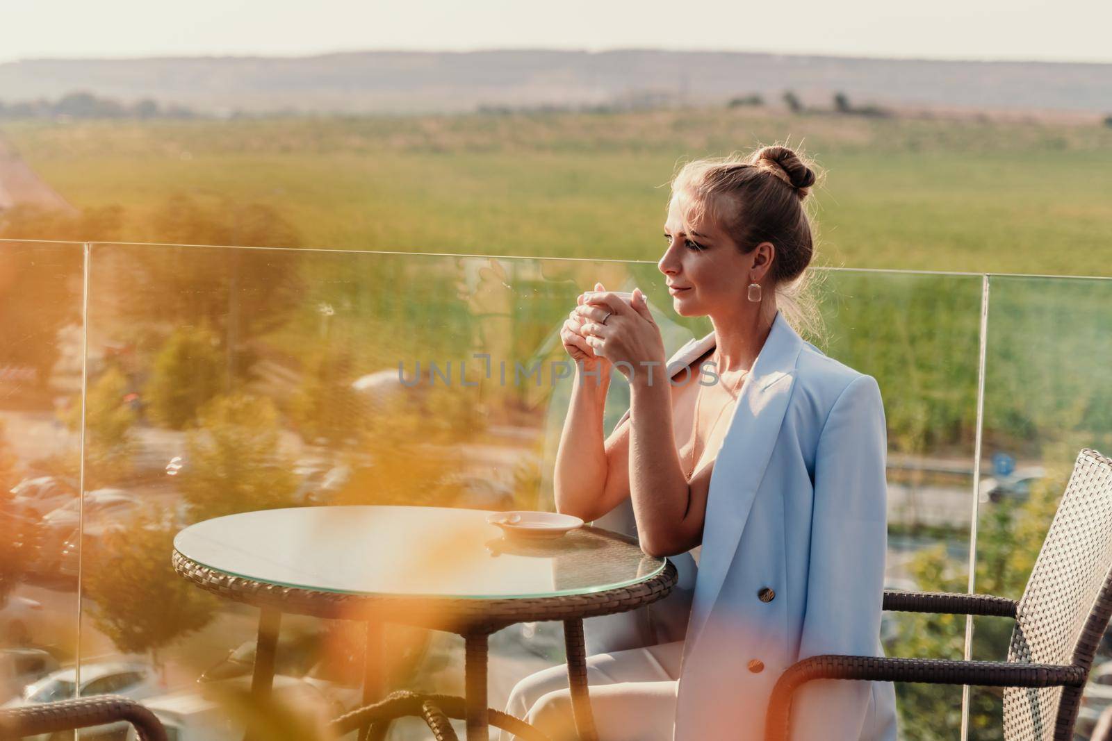 A middle-aged woman sits in a street cafe overlooking the mountains at sunset. She is dressed in a blue jacket and drinks coffee admiring the nature. Travel and vacation concept. by Matiunina