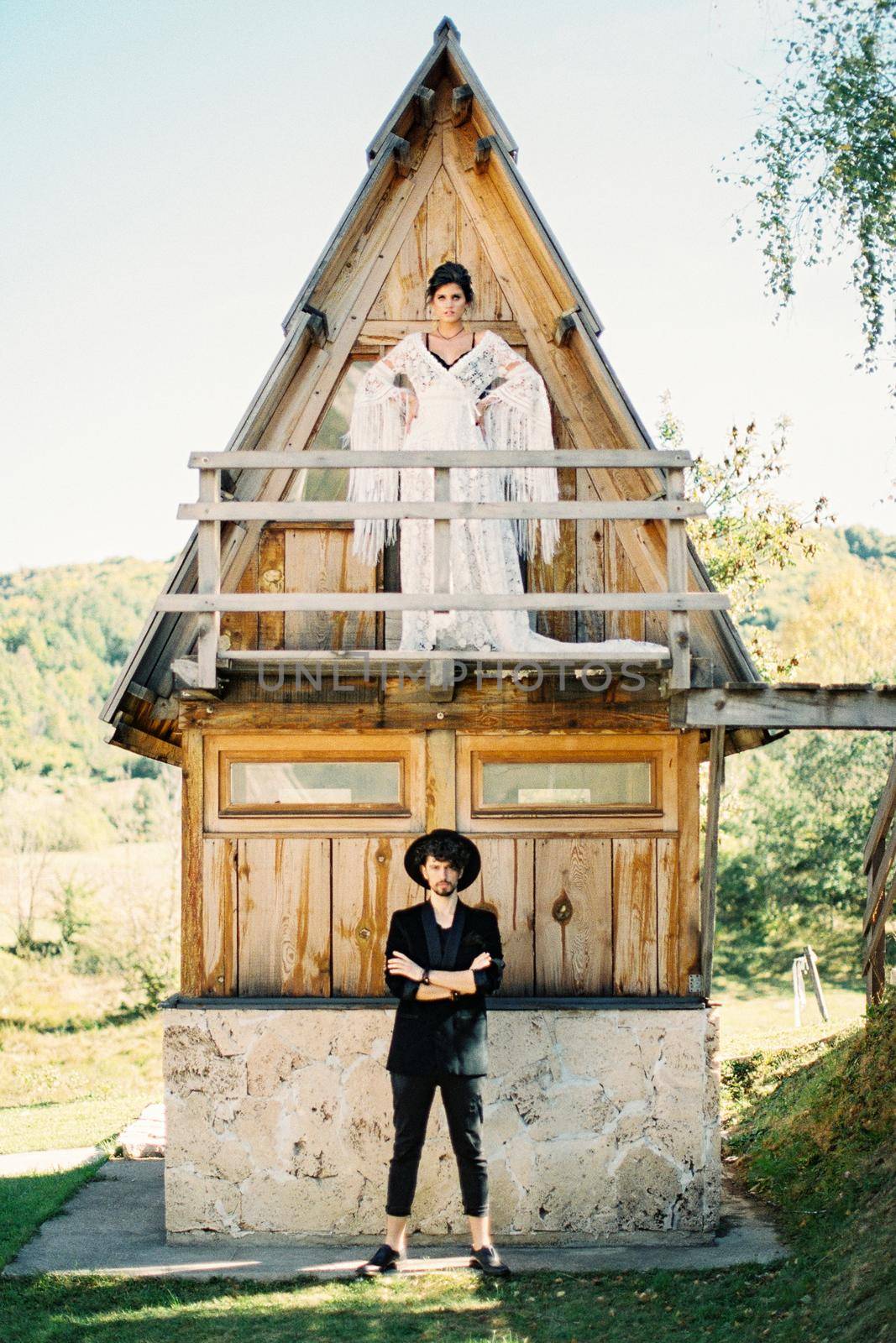 Bride stands on the balcony of a triangular house, above the groom standing on the pavement. High quality photo
