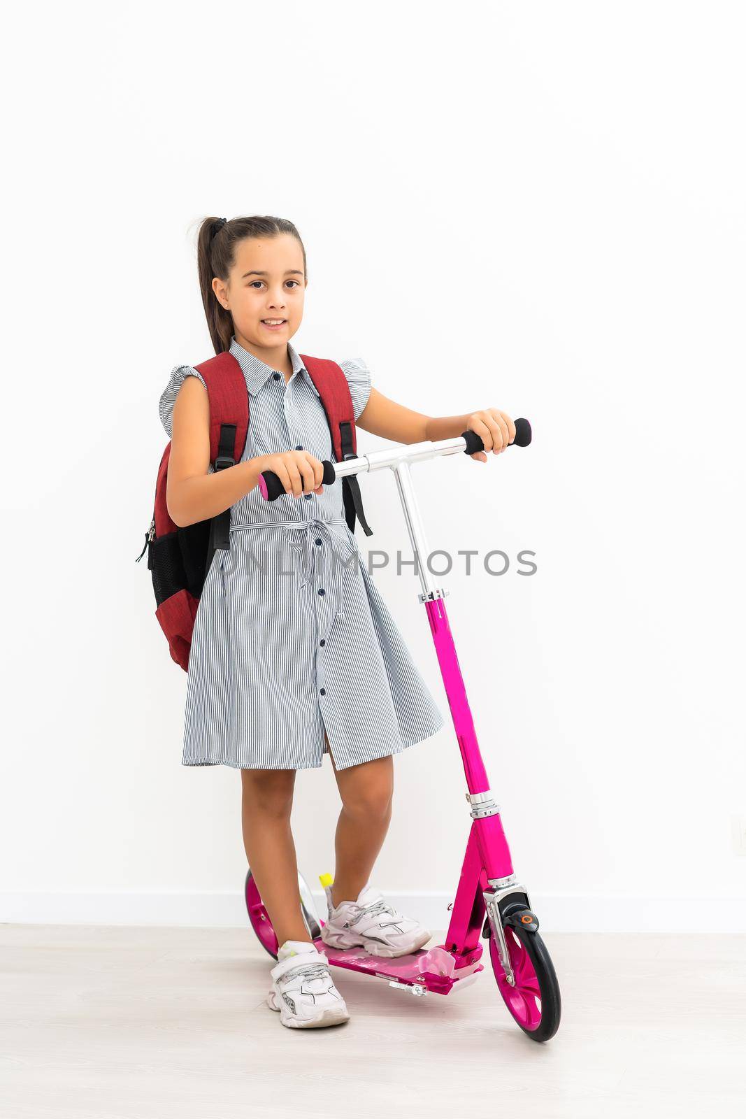 Full length profile shot of a schoolgirl with a backpack riding a scooter isolated on white background.