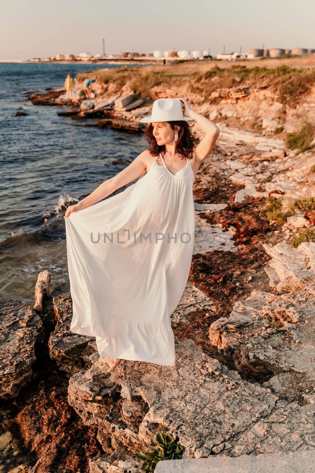 A woman in a white dress and hat is standing on the beach enjoying the sea. Happy summer holidays.