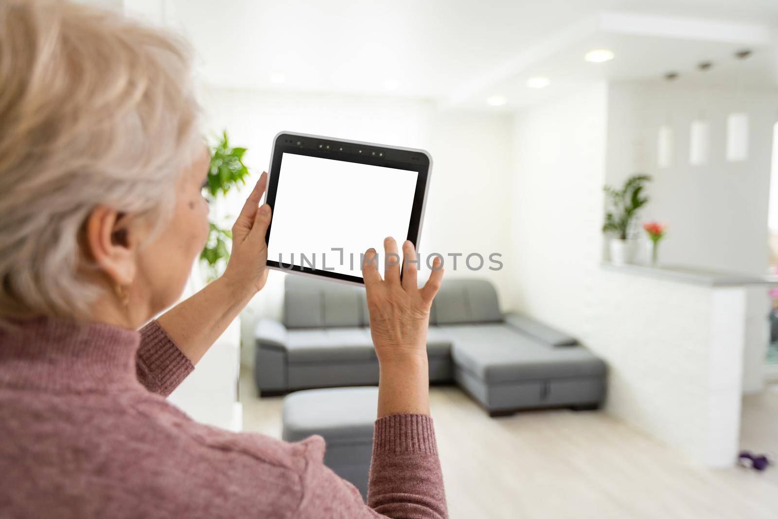 Senior woman at home standing at kitchen holding digital tablet controlling smart home system back view checking cameras close-up.
