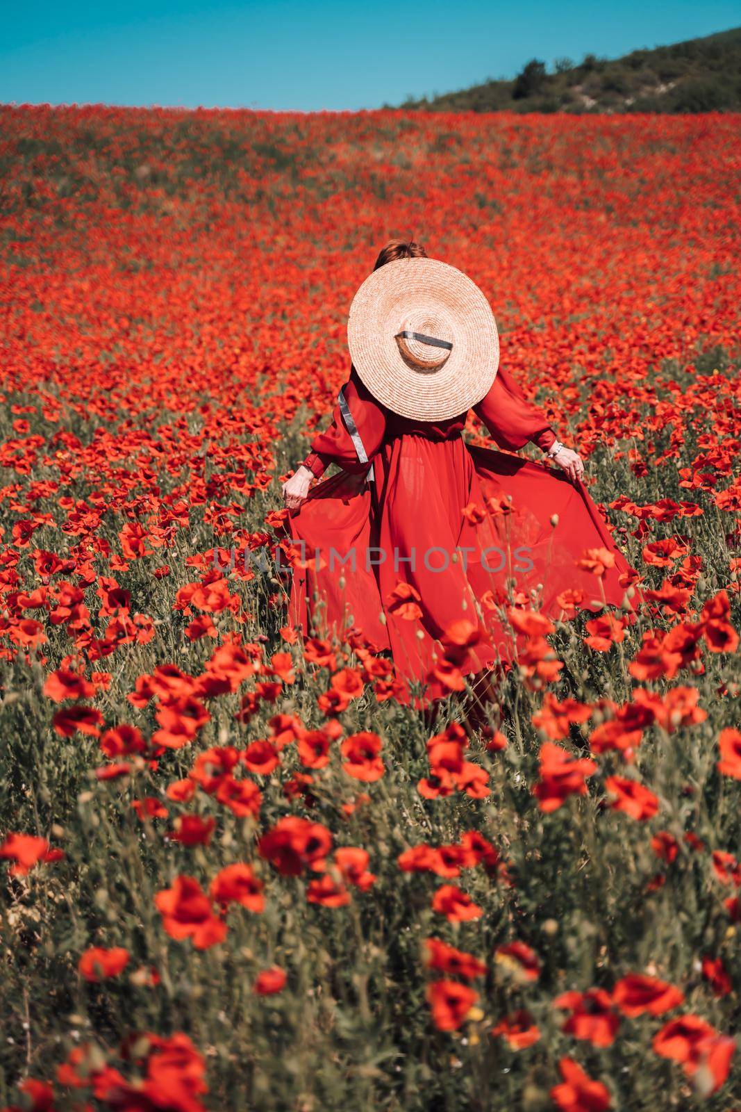 Young woman stands with her back in a long red dress and hat, posing on a large field of red poppies by Matiunina